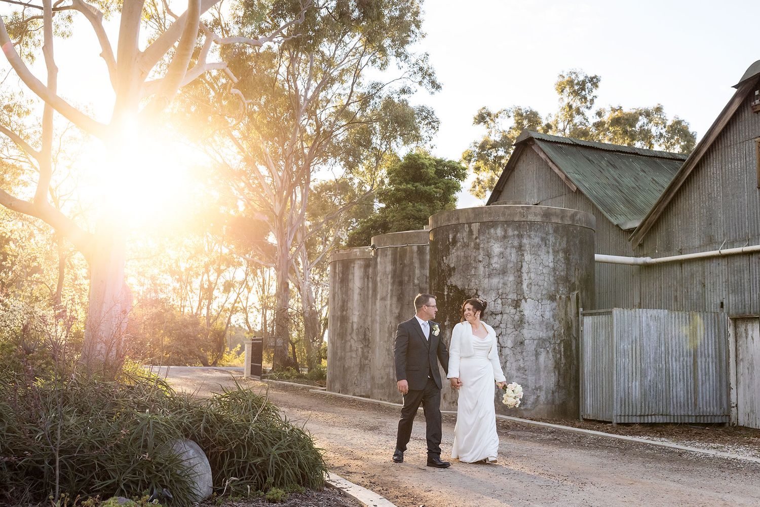 A Bride and Groom walk hand in hand beside an old shed at Buller Wines at Sunset.