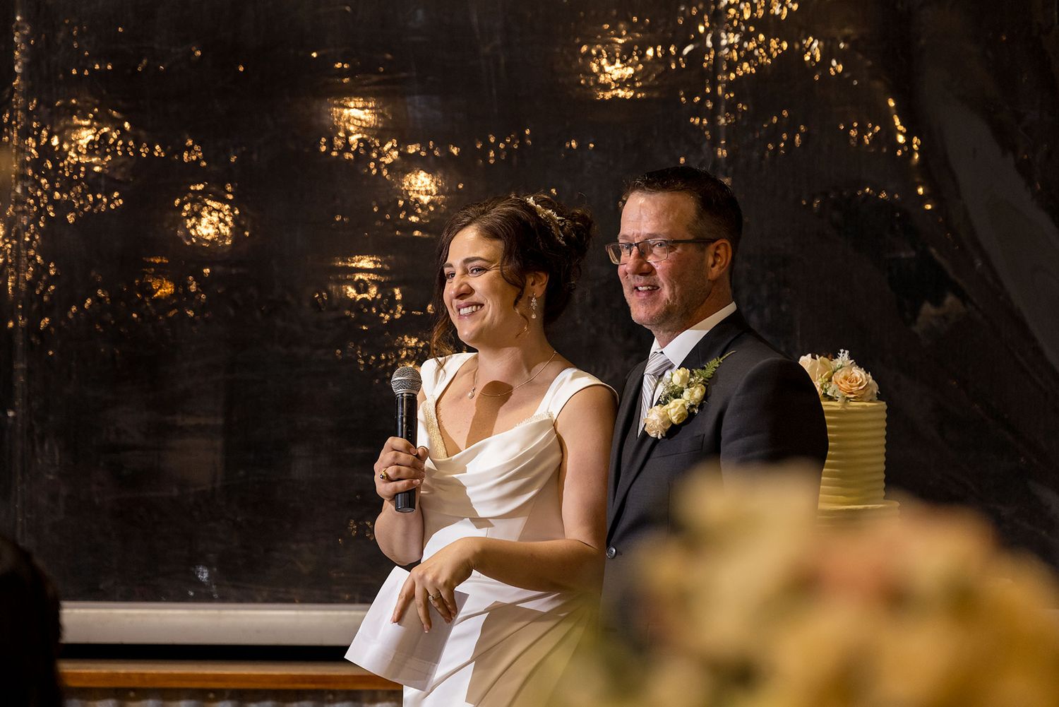A joyful wedding couple shares a laugh together during their speech at their reception with glowing lights in the background at Buller Wines