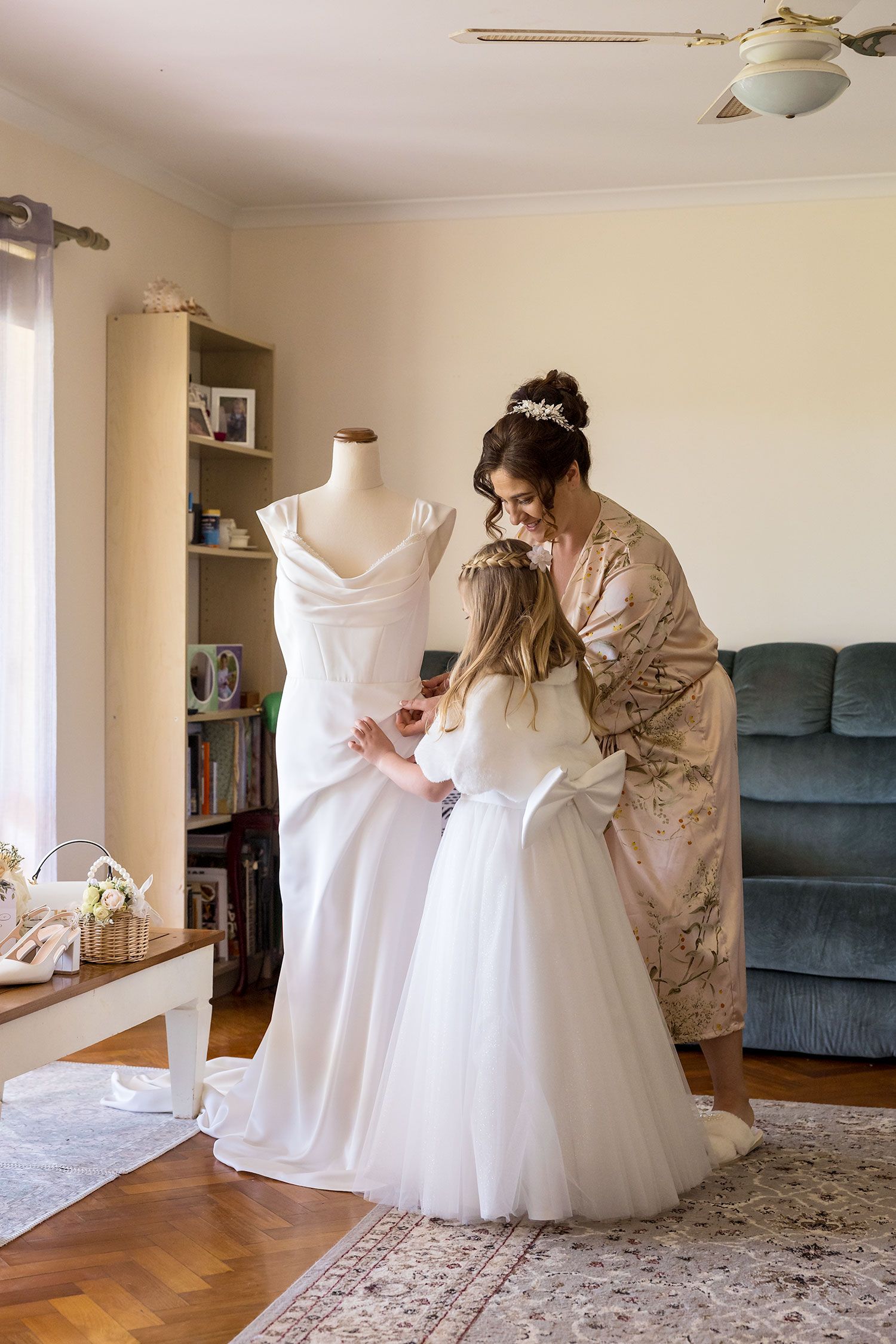 A photo showing a Bride with her daughter adjusting a white wedding dress on a mannequin  in a living room setting.