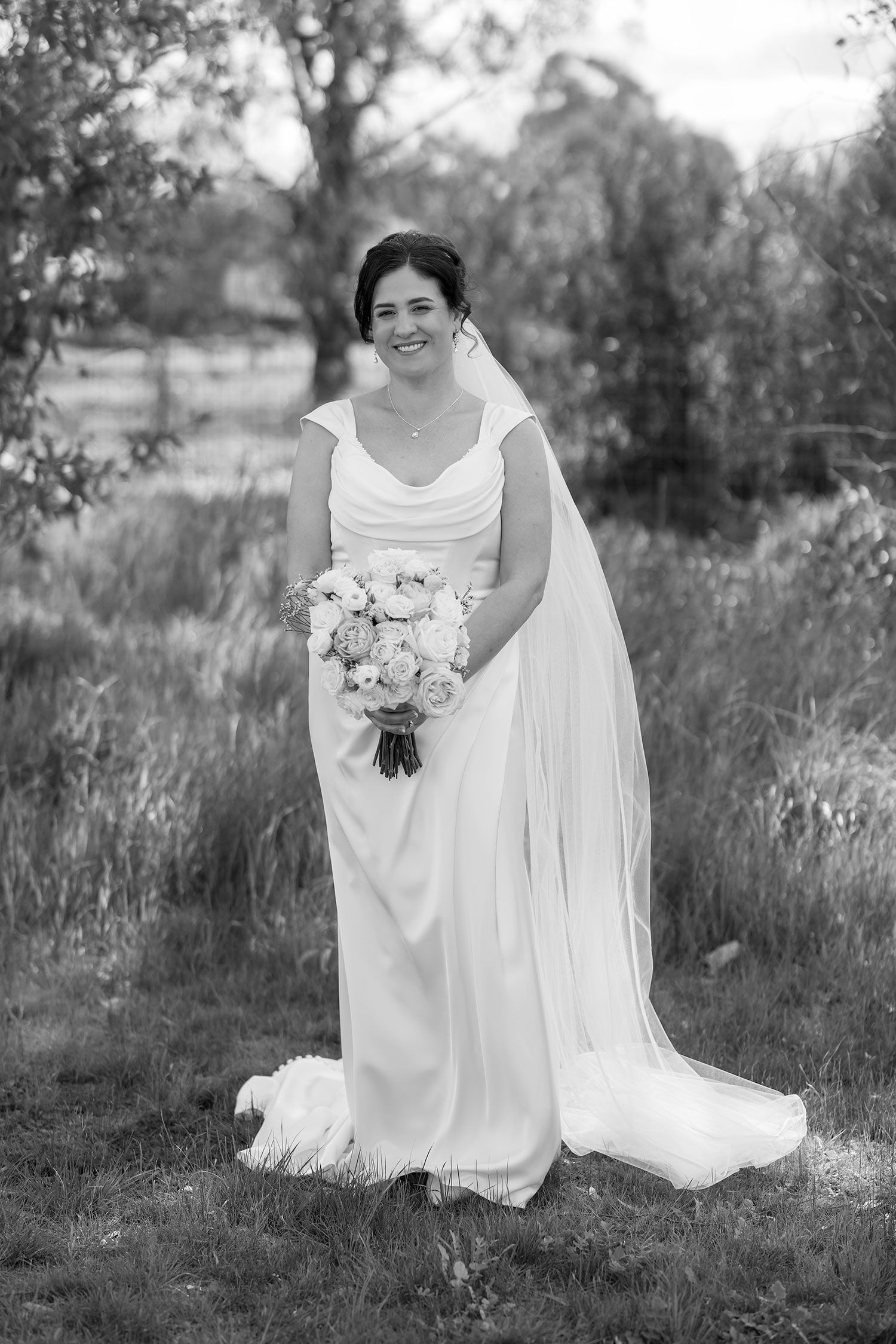 A bride in a flowing white dress sits gracefully in a meadow with tall grass in black and white photography.
