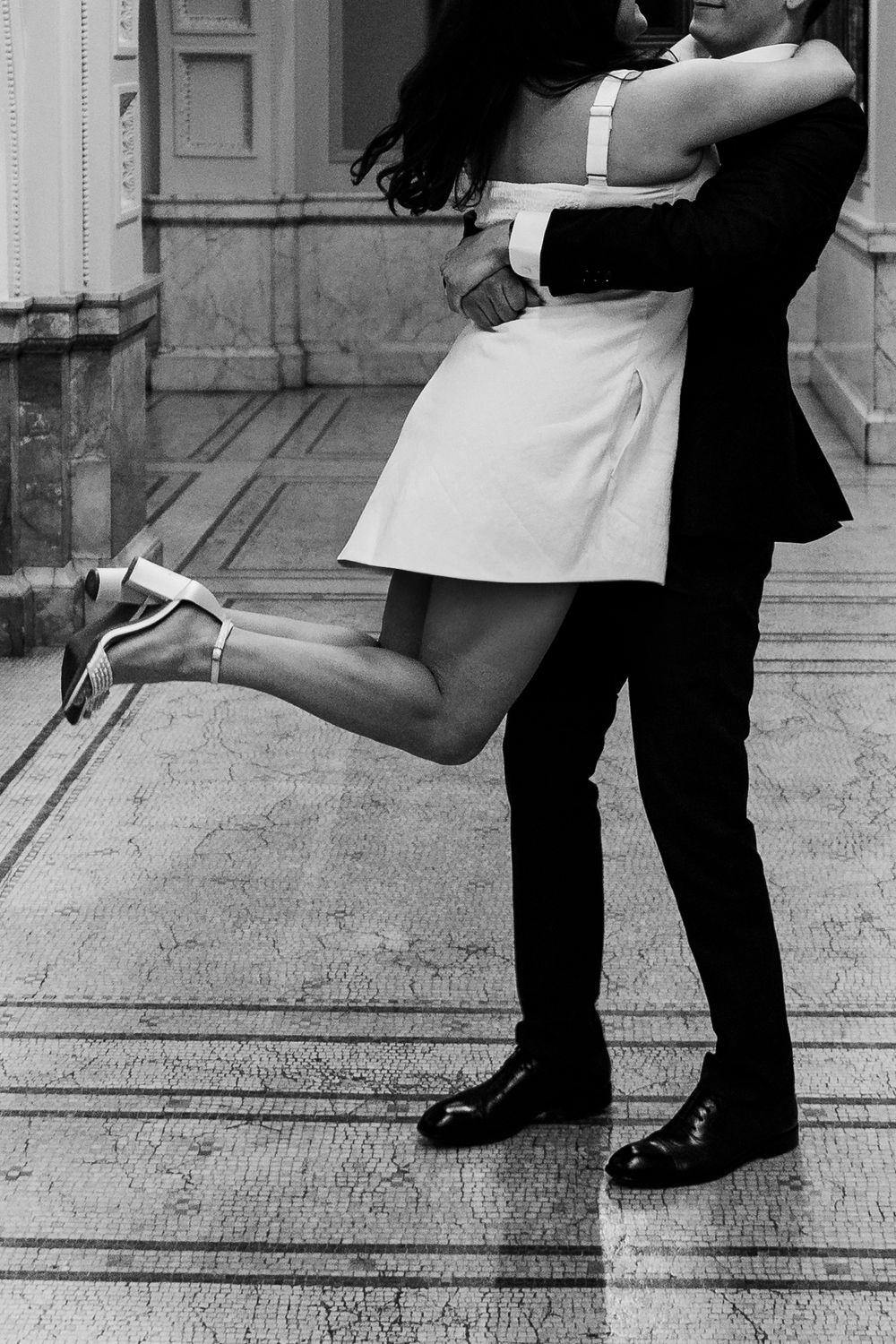 A couple embraces in formal attire, with the woman's feet lifted off the ground in a playful moment.