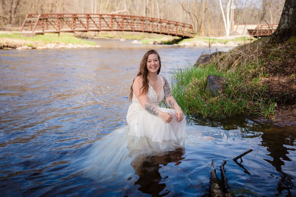 A white wedding dress flows in the water near a rustic bridge over a river during a bridal photo session.