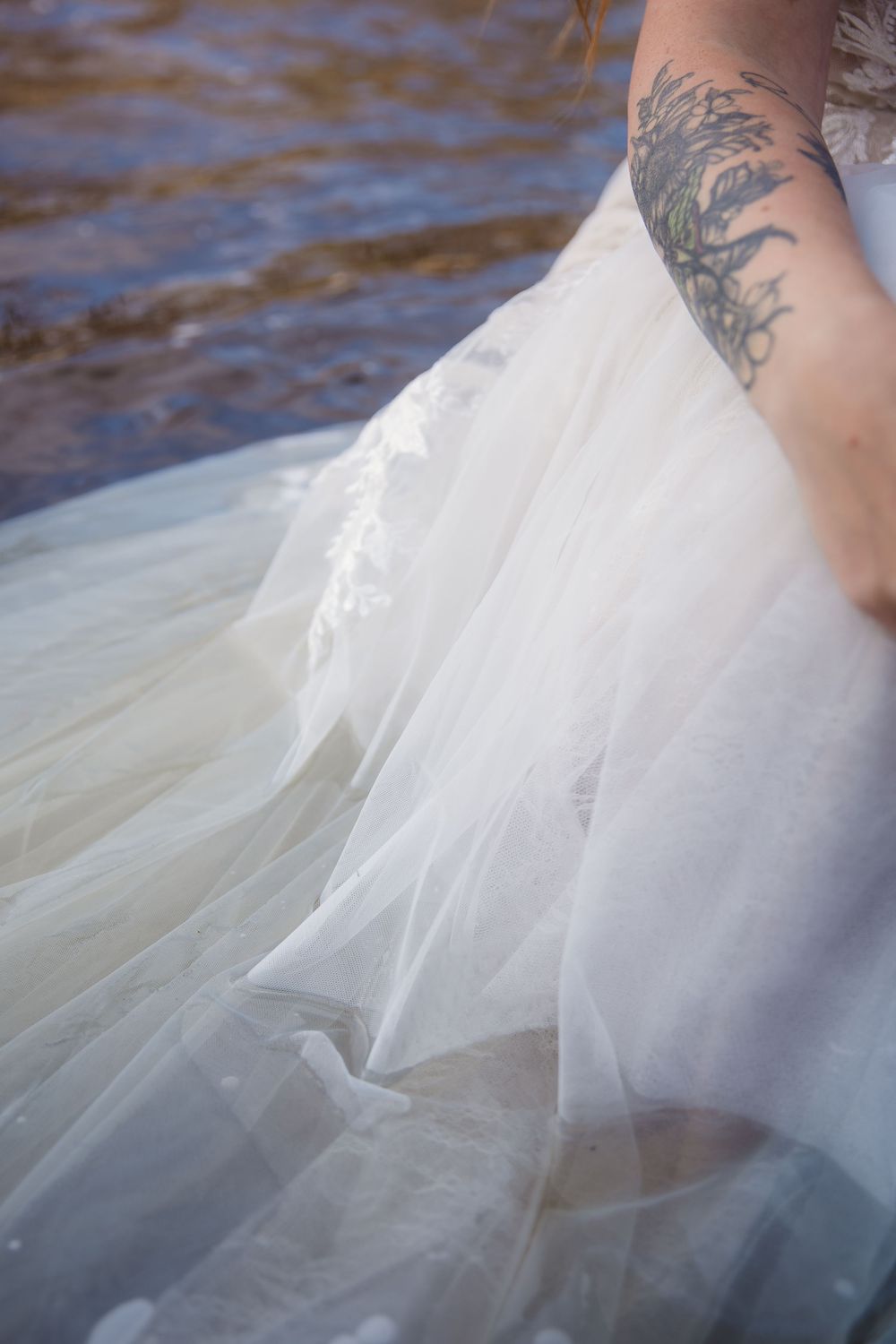 Close-up of flowing white dress fabric and tattooed arm in water.