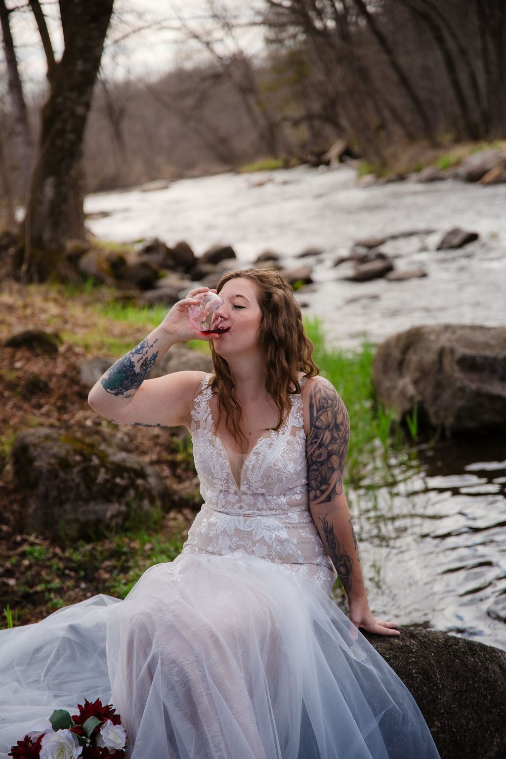 Woman in wedding dress drinking wine by riverside with tattoos visible.