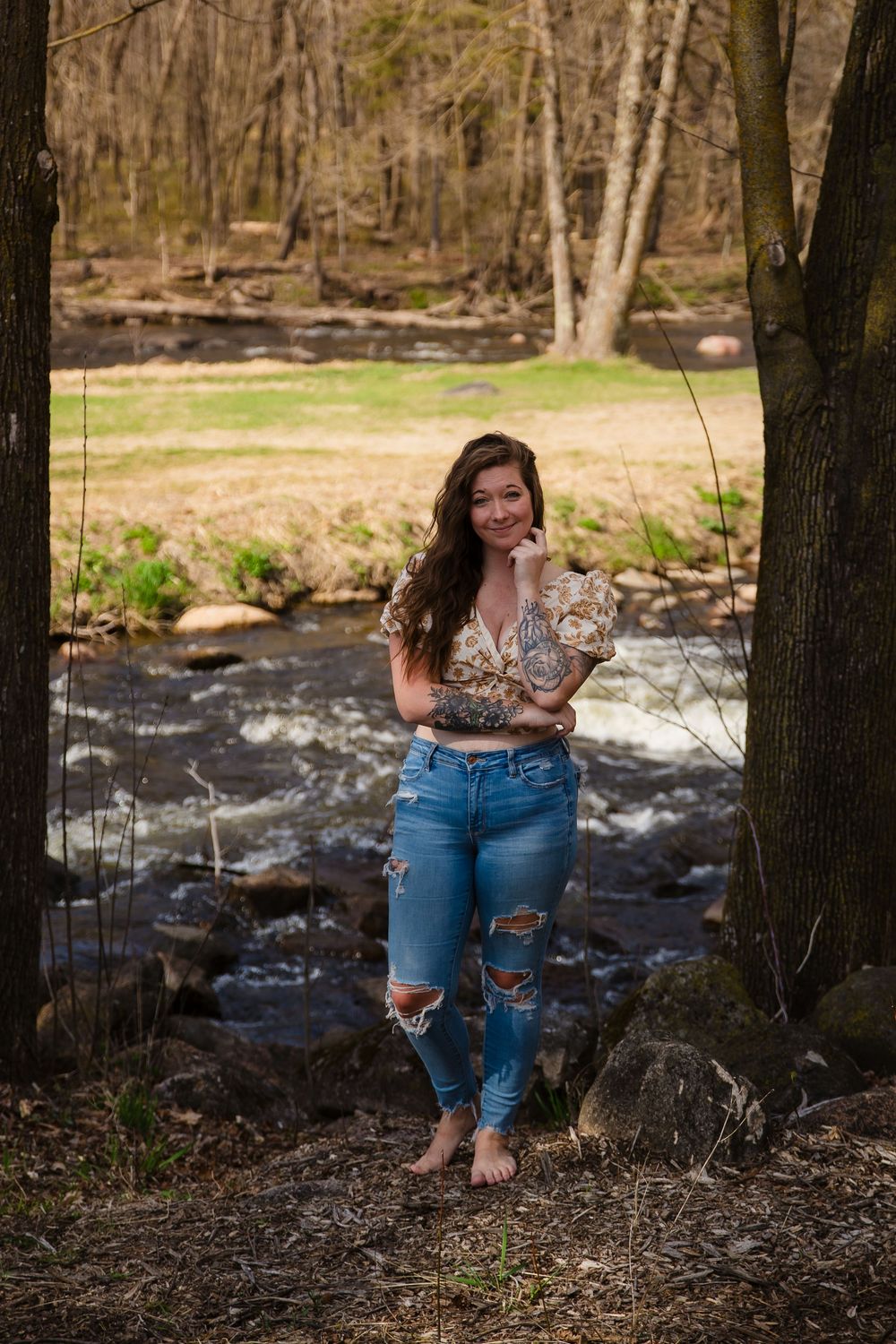 Woman in ripped jeans posing by rushing stream in wooded setting.