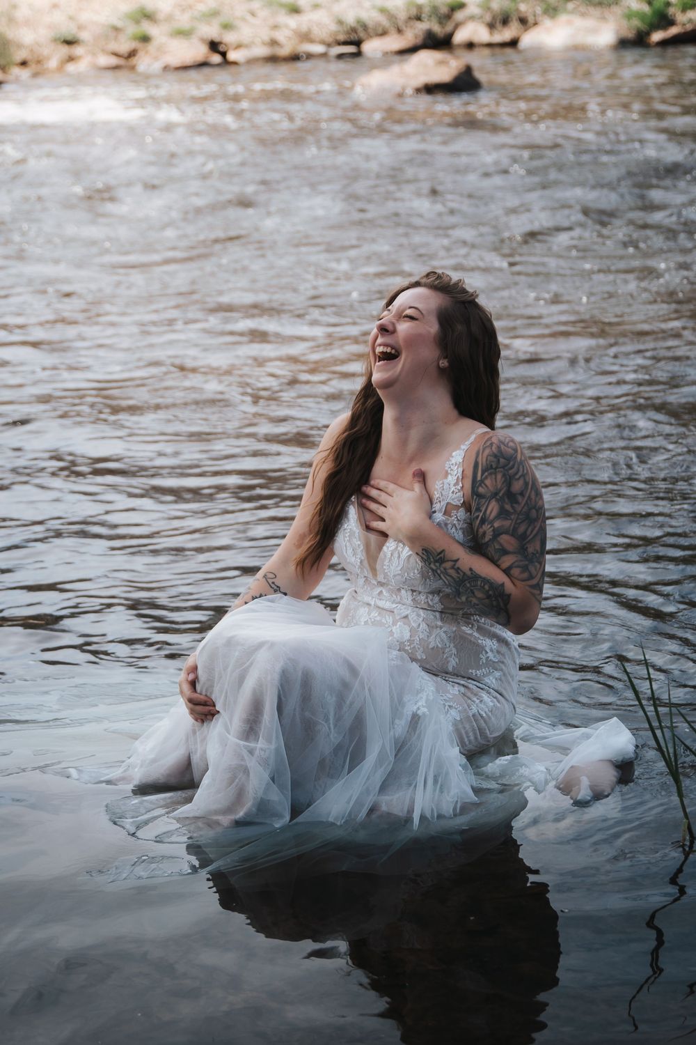 Woman in white dress laughing joyfully while sitting in flowing river.
