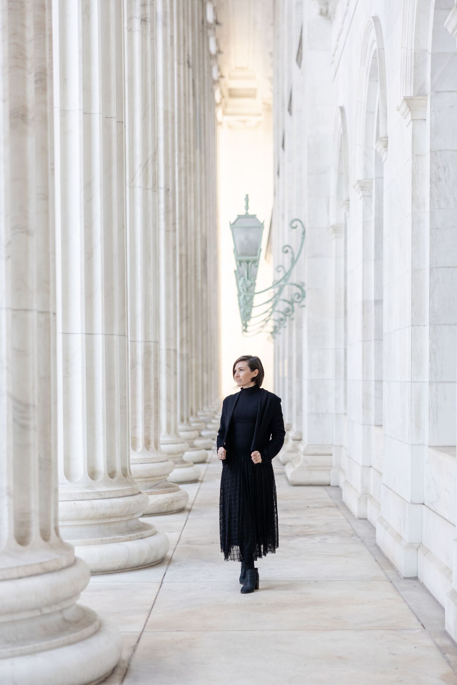 A person in a black dress walks through a colonnade of white pillars.