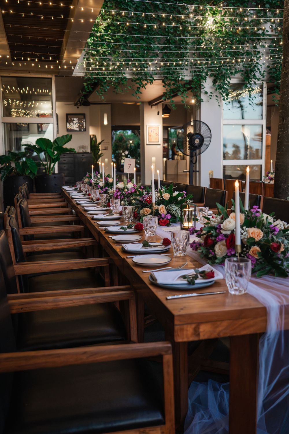 Long wooden table decorated with floral arrangements and delicate blue runners under romantic string lights at a wedding