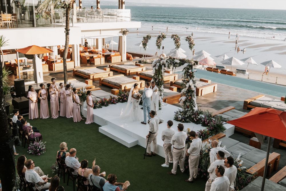 An elegant beachfront wedding ceremony setup with white chairs and floral arrangements overlooking the ocean with wedding guests