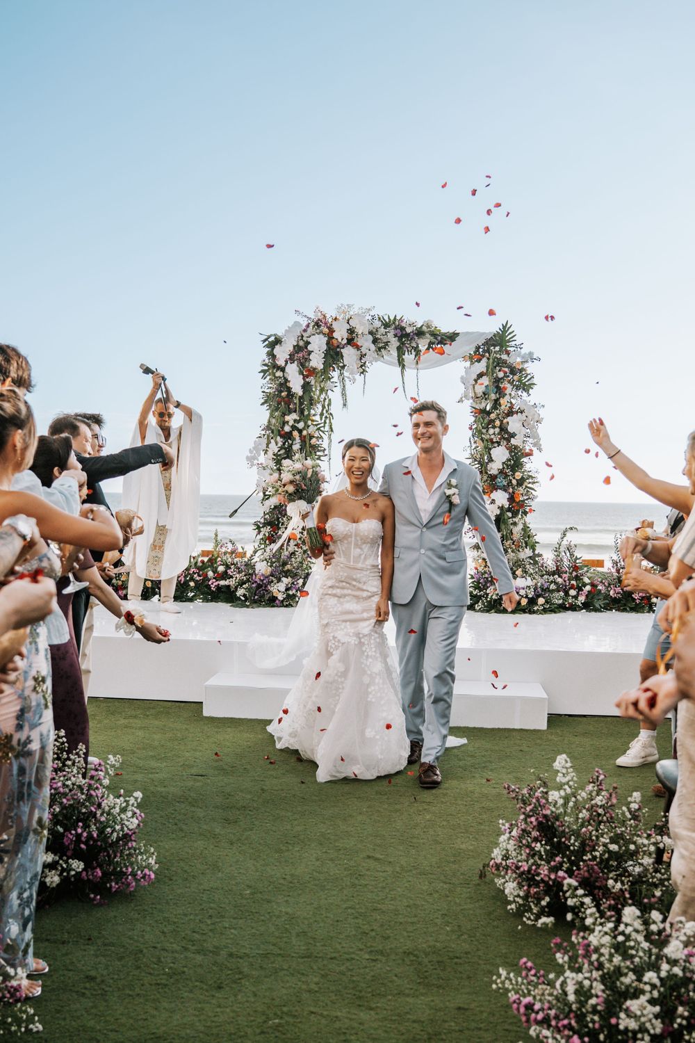 Joyful wedding celebration on a grassy lawn with floral arch decorations and guests throwing flower petals at newly married couple