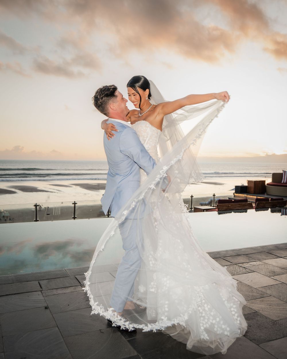 A couple dances together on a beachfront patio during sunset with a pool reflecting dramatic clouds in the background.