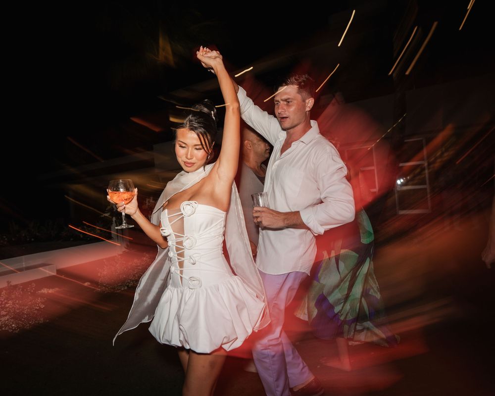 A couple in wedding attire dances energetically in a dimly lit reception venue with red ambient lighting.