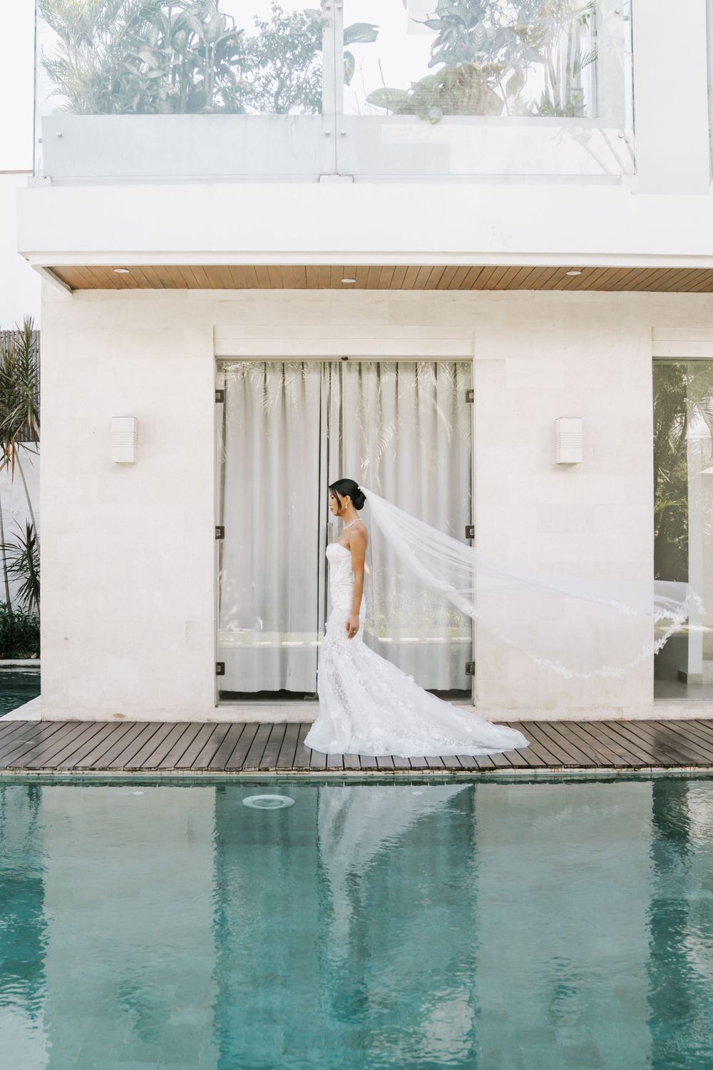 Elegant bride in a flowing white gown walking along a poolside with white curtains and modern architecture