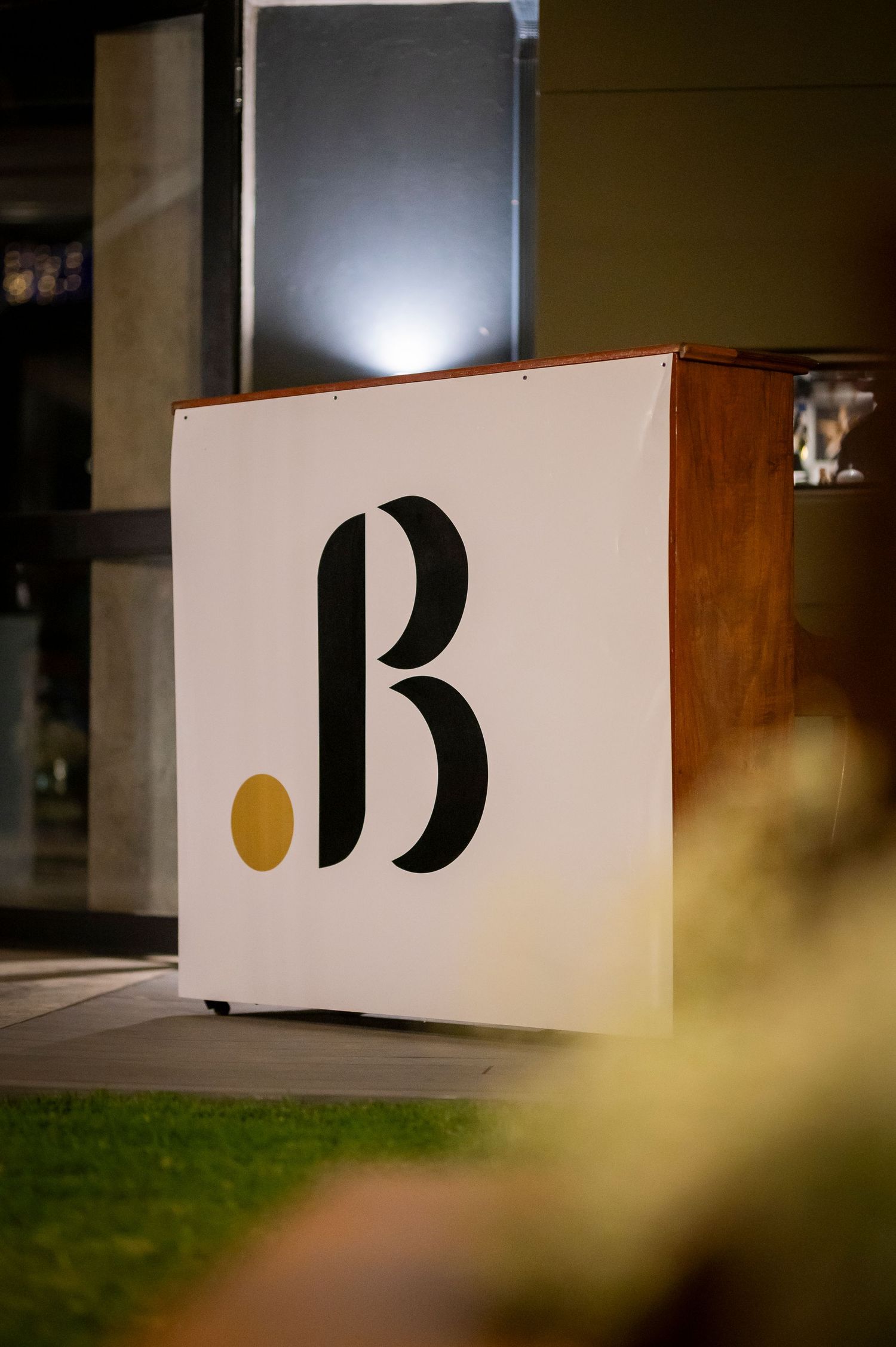 A minimalist white box sign displays a large black letter 'B' with a small yellow dot accent against a wooden surface.