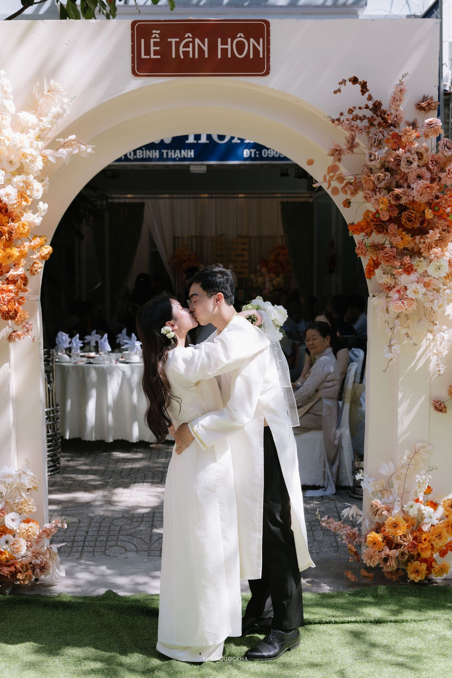 Vietnamese bride and groom kissing under arch decorated with orange flowers at Le Tan Hon wedding ceremony.