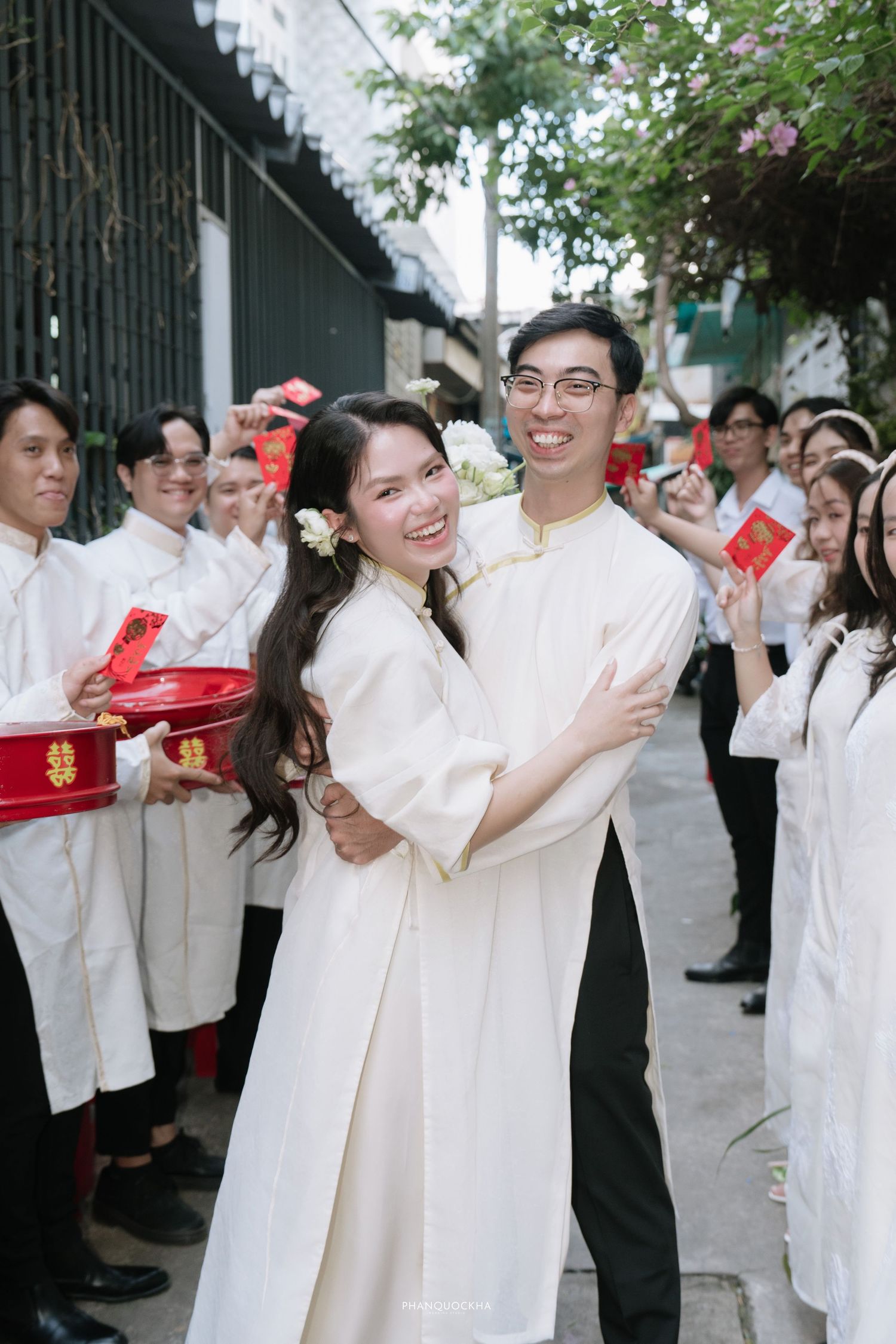Smiling Vietnamese bride and groom embrace in white ao dai during traditional wedding ceremony with guests.