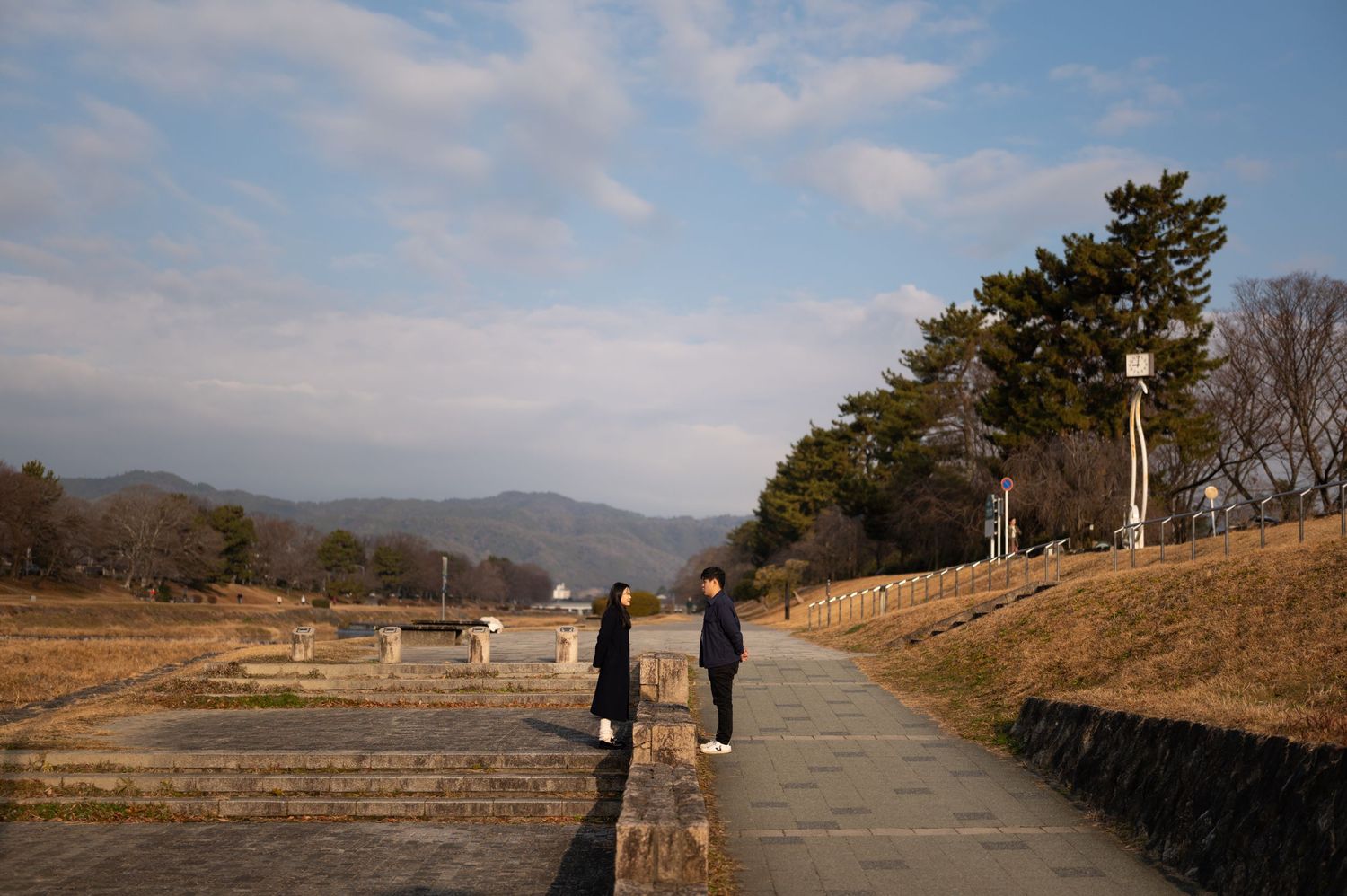 Couple walking along a stone path near historic ruins with pine trees and mountains in background.