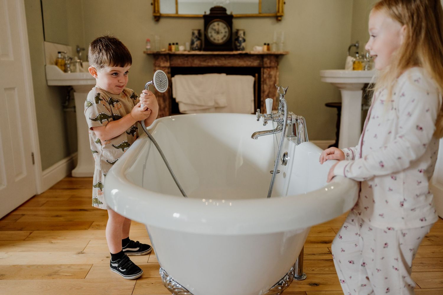A child in white clothing leans over a freestanding bathtub in a bright, minimal bathroom setting.