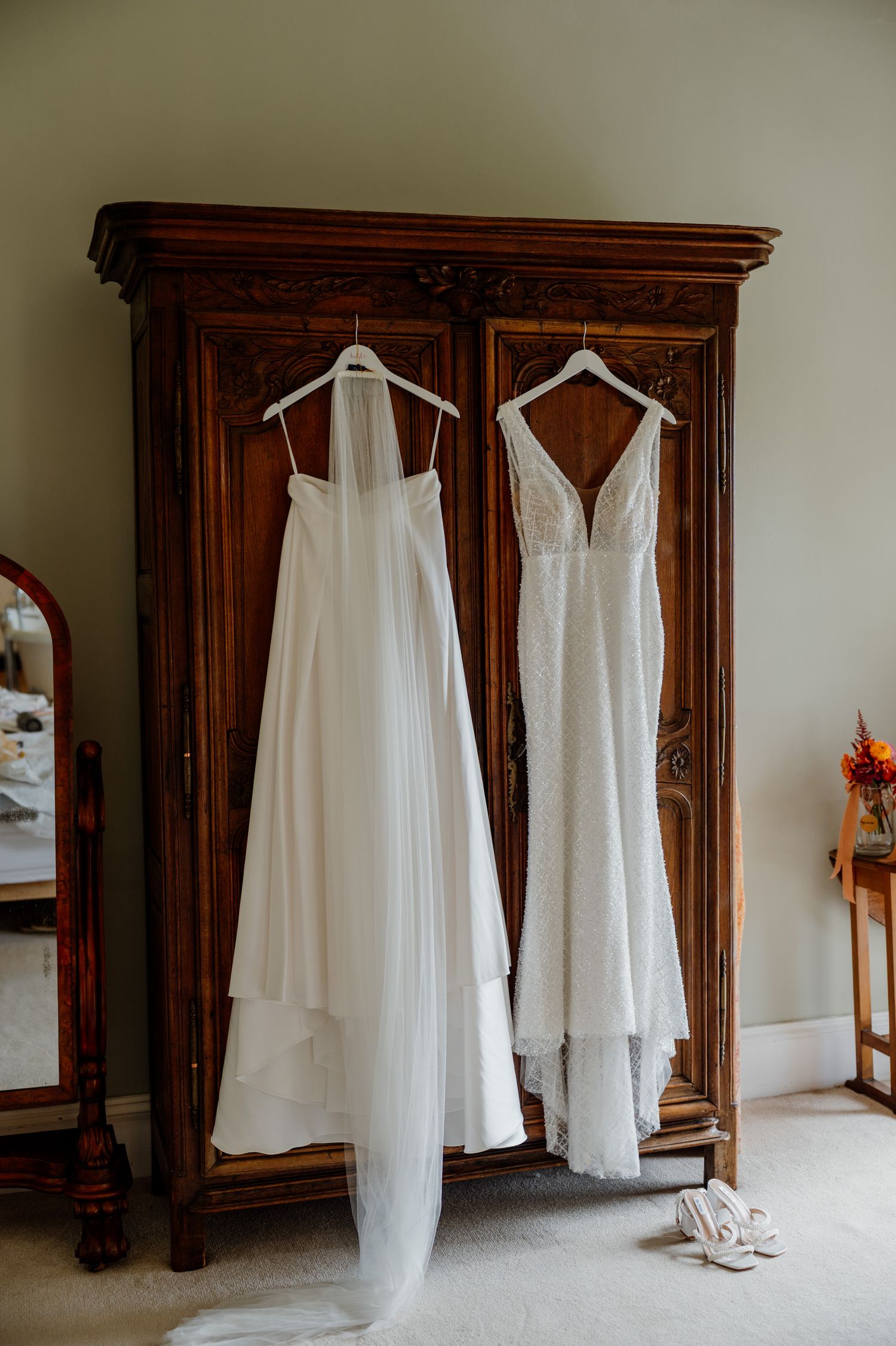 White wedding dresses and veils hanging against ornate wooden wardrobes in a classic dressing room setting.