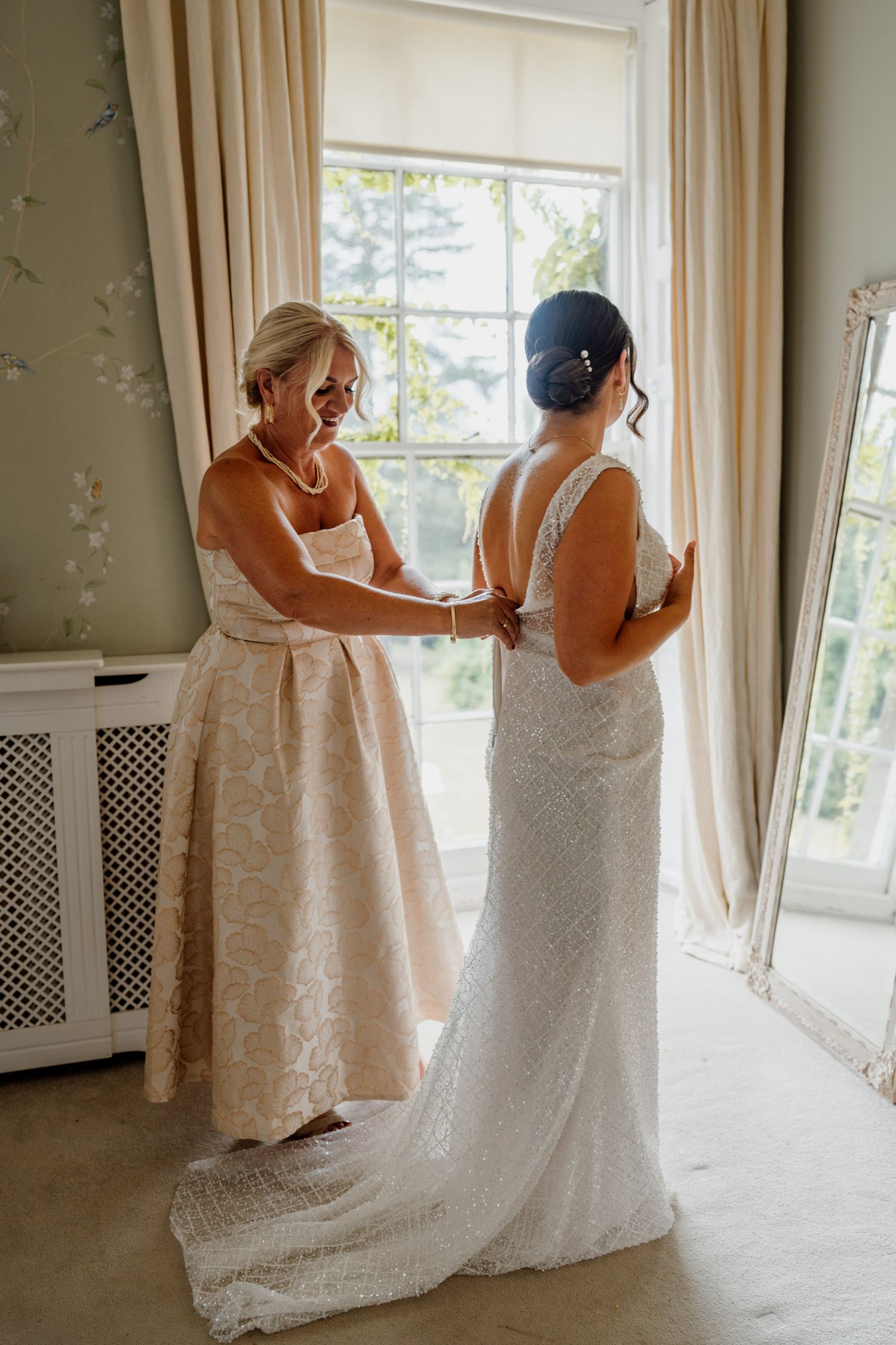 Wedding dress preparation scene in a sunlit room with cream curtains and floral wallpaper.