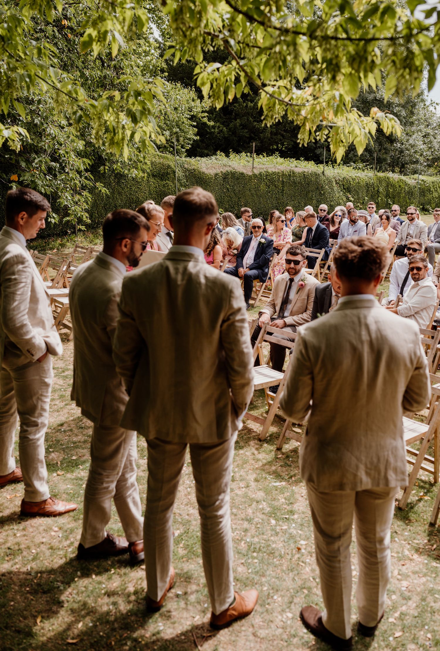 Wedding party in tan suits gathered outside under green trees.