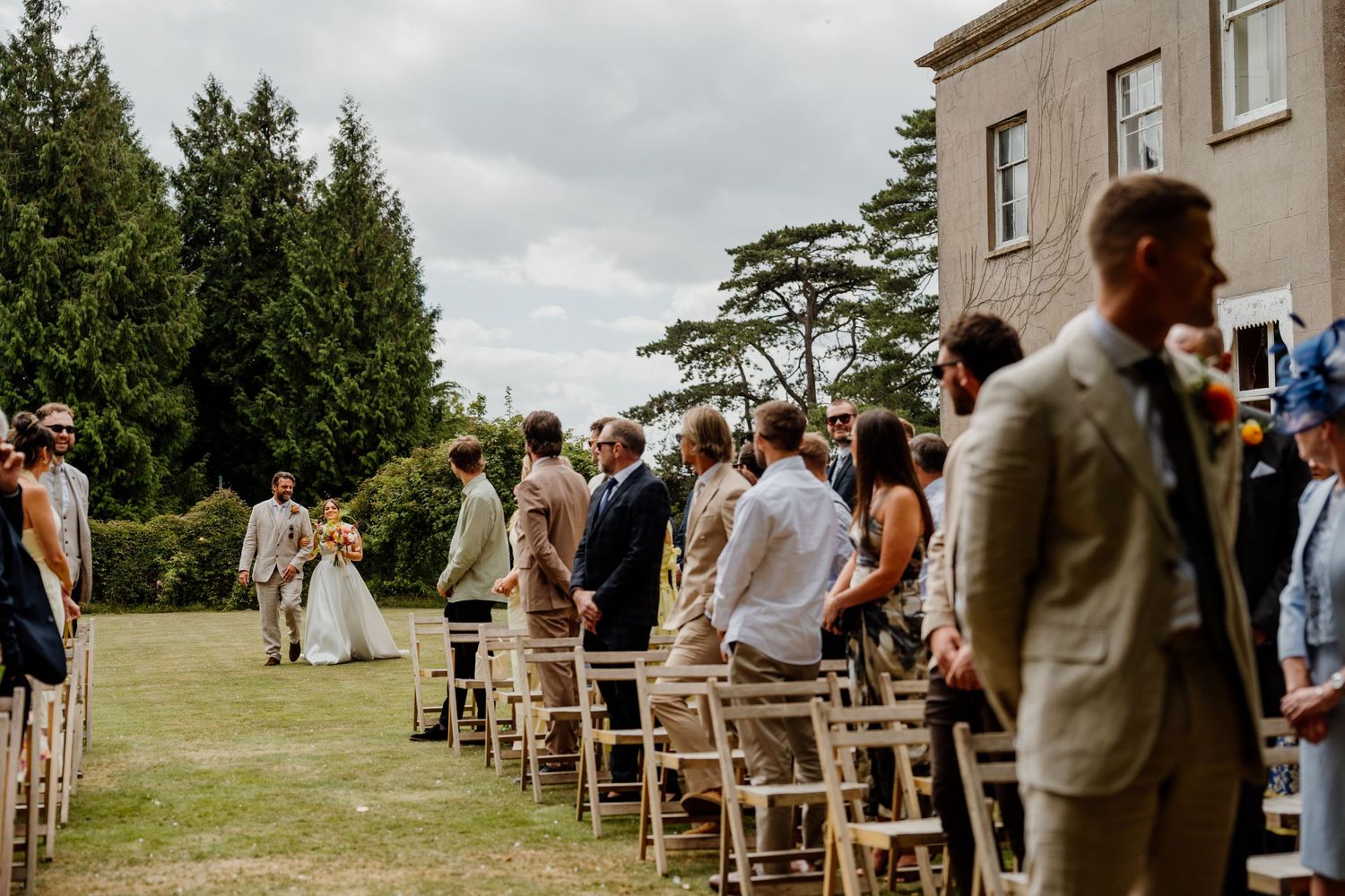 Outdoor wedding ceremony setup with wooden chairs facing a stone building under cloudy skies.