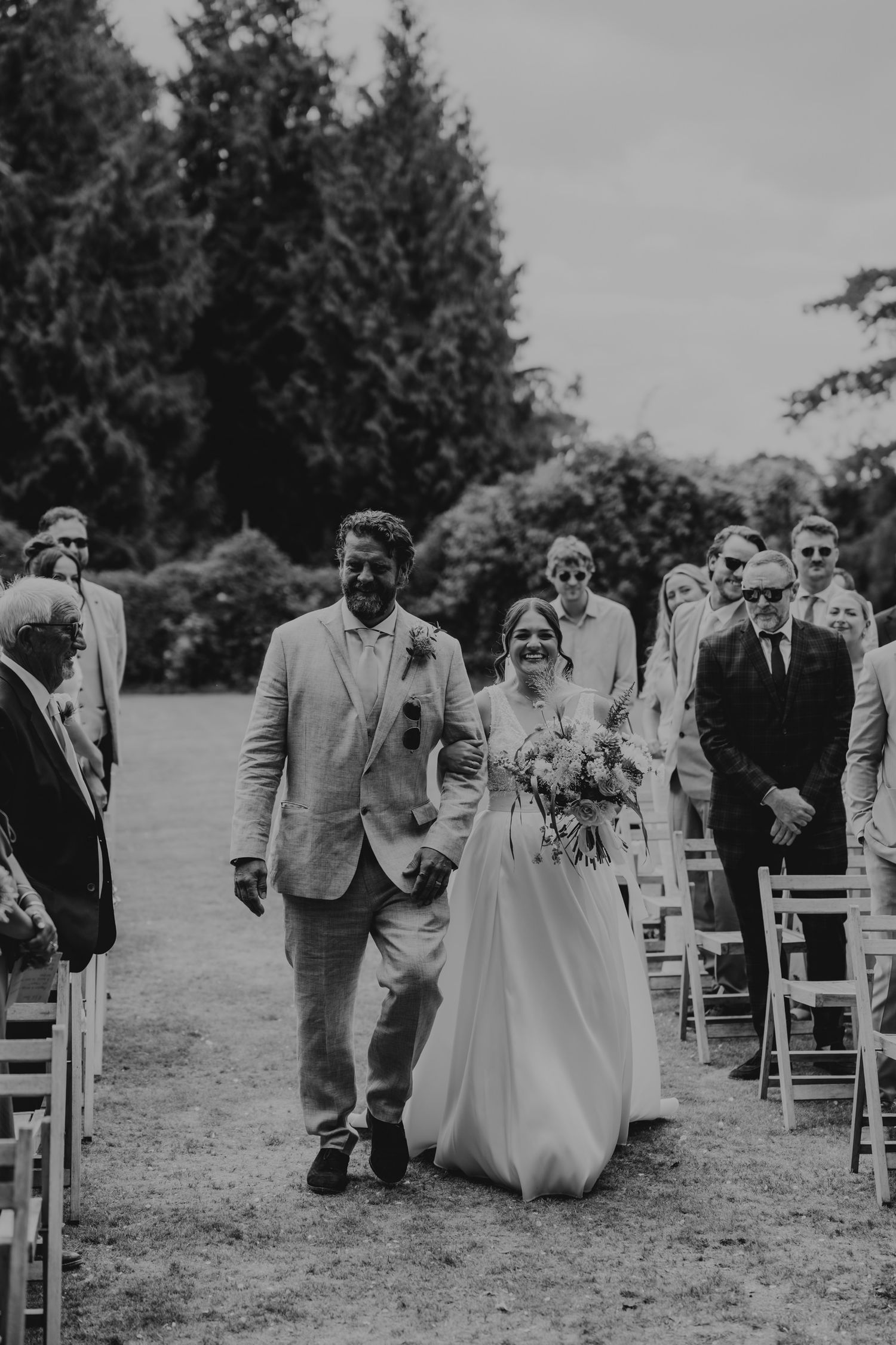 Wedding guests walking down an aisle of white chairs on a grassy lawn with evergreen trees in the background.