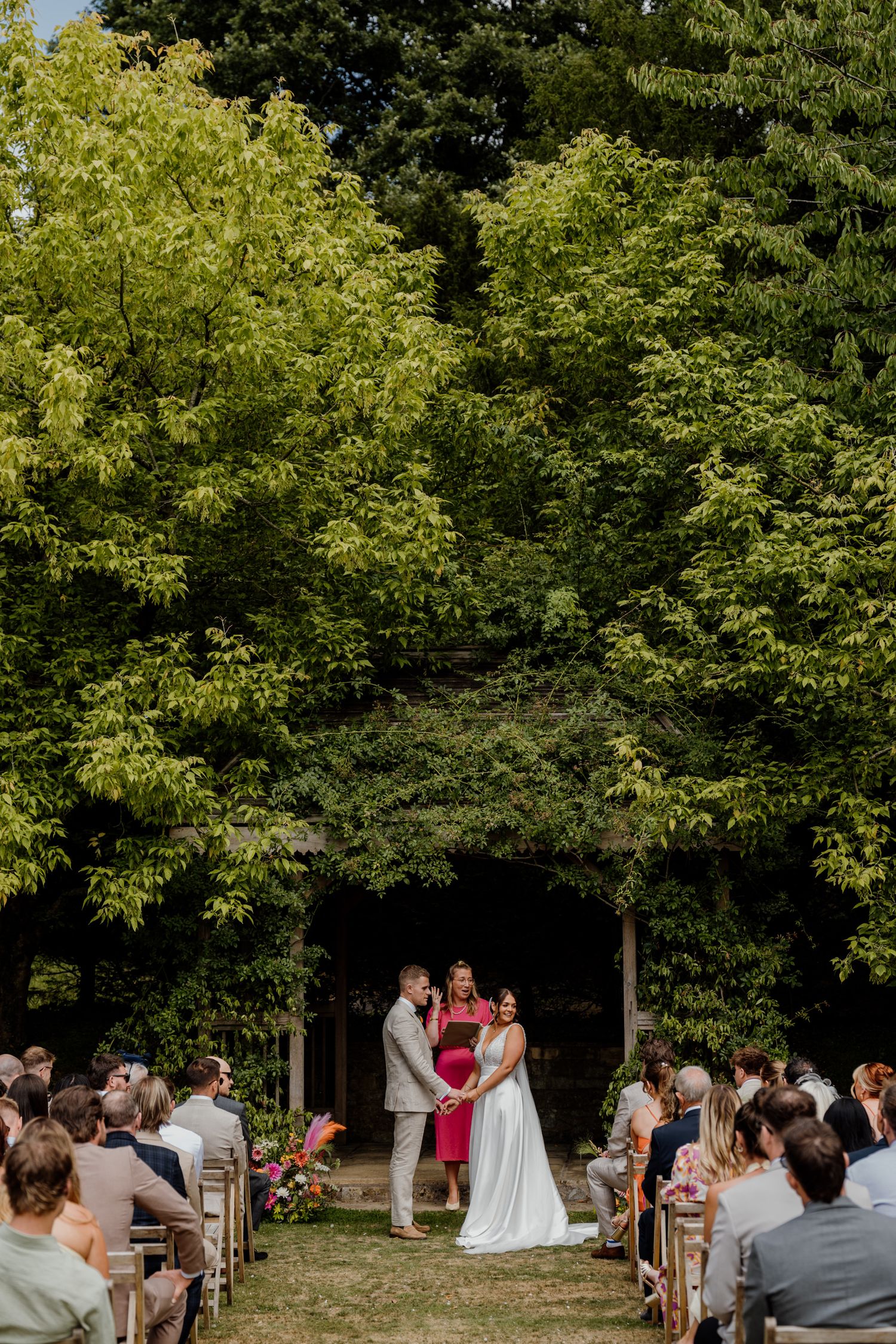 Outdoor wedding ceremony in a garden setting surrounded by lush trees.