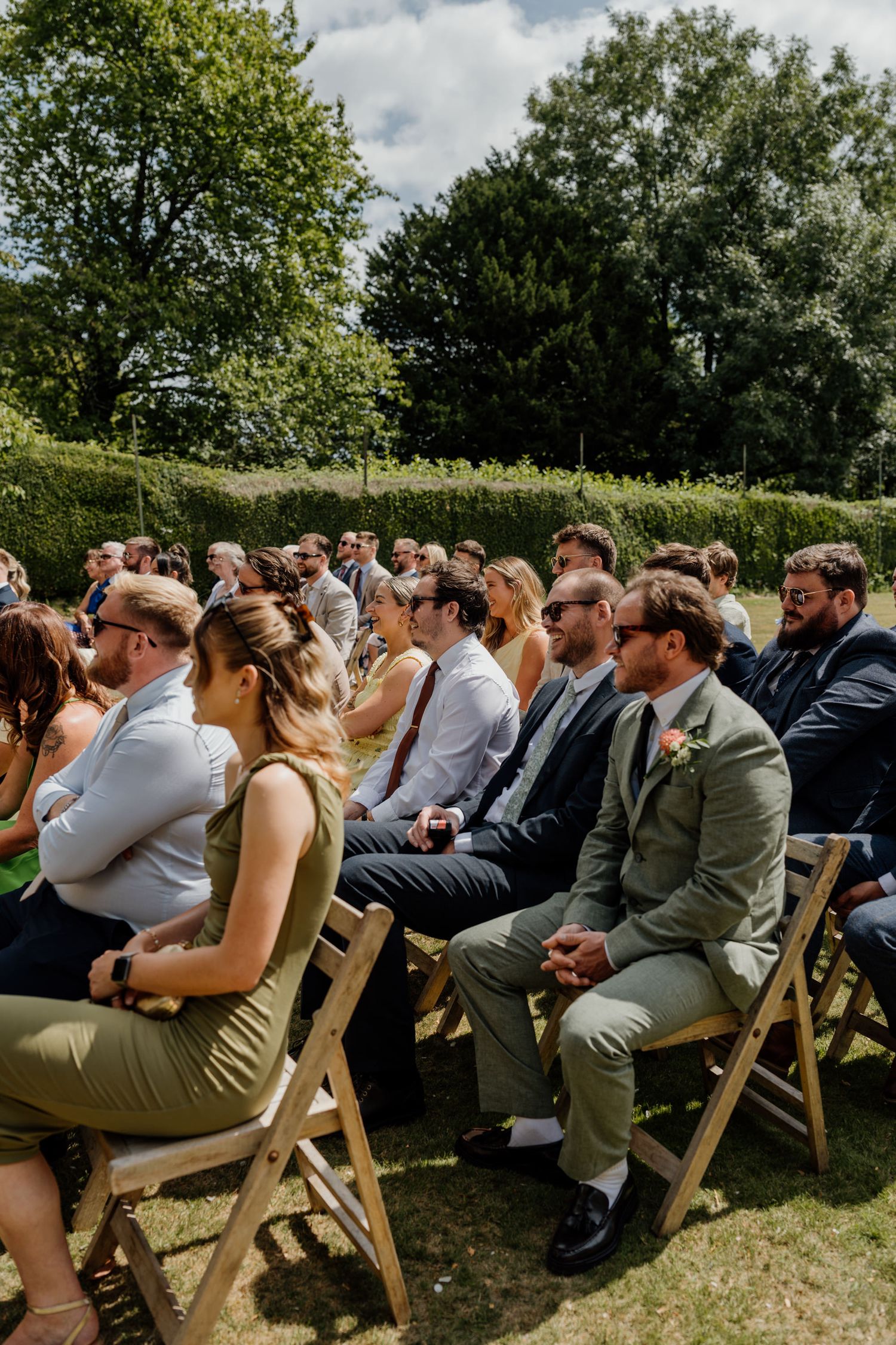 Wedding guests seated in wooden chairs at an outdoor ceremony under green trees.