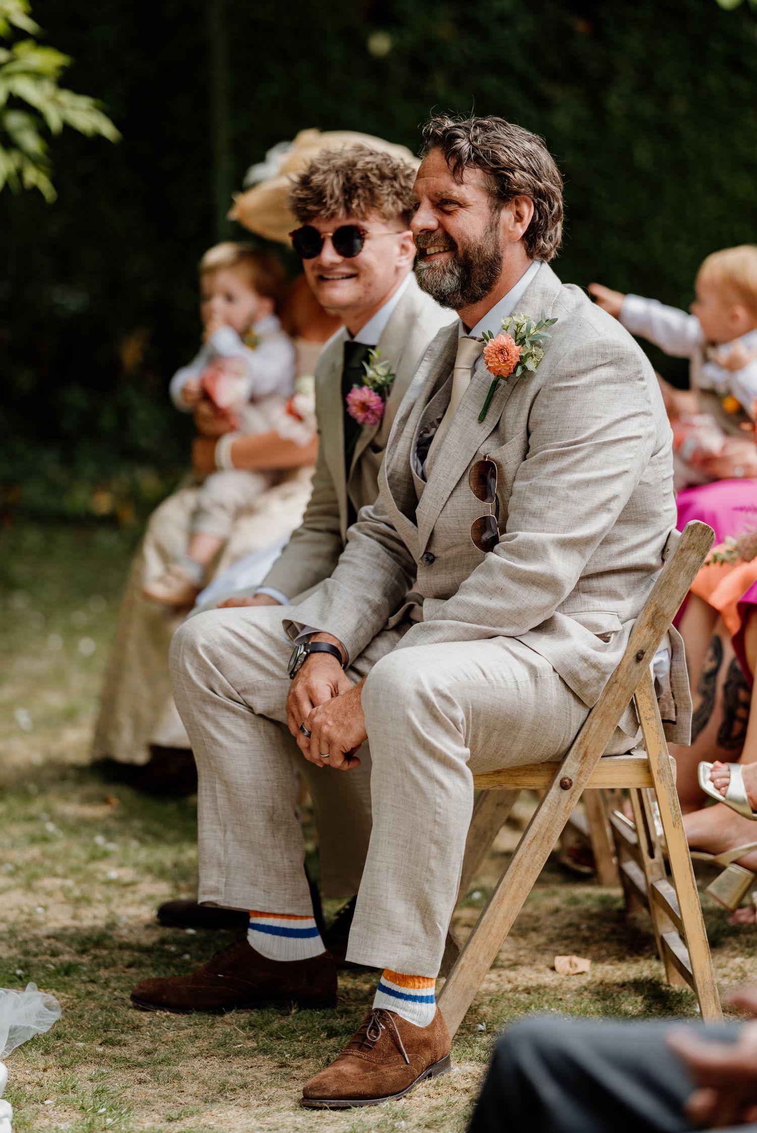Guests in light beige suits sitting on wooden chairs during an outdoor event.