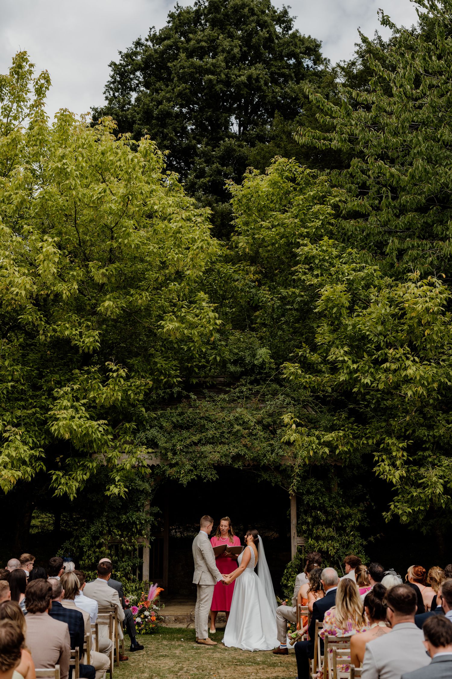 An outdoor wedding ceremony with guests seated under a canopy of green trees on a sunny day.