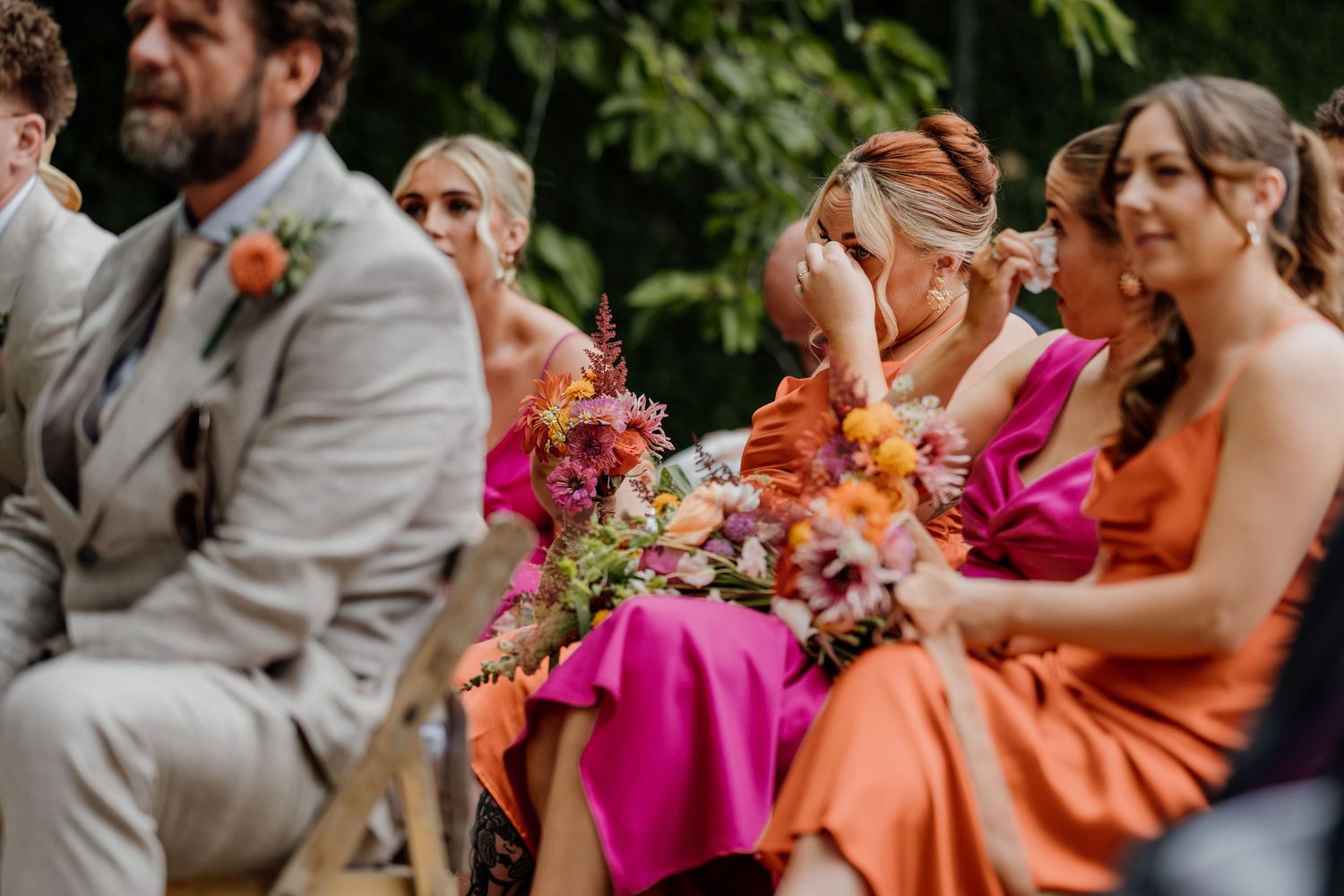 Wedding party in bright pink and orange dresses reacting with laughter during outdoor celebration.