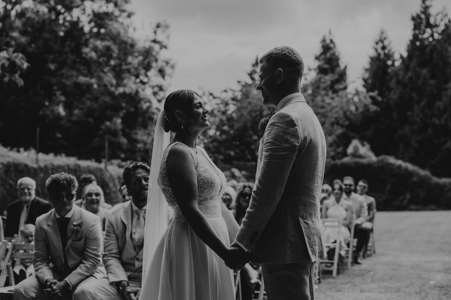 Wedding guests stand in rows during an outdoor ceremony beneath tall trees.