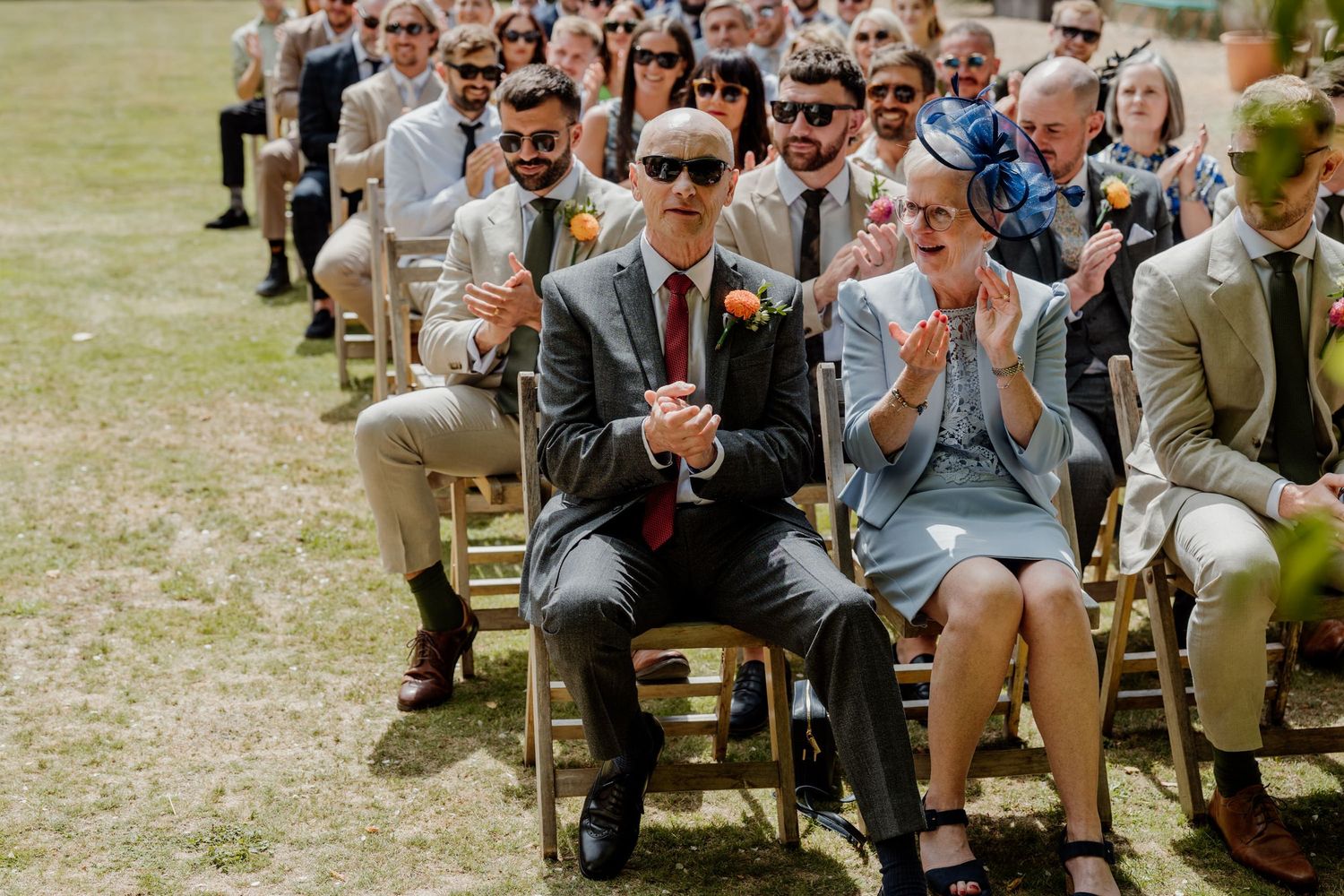 Wedding guests seated in rows of wooden chairs on a grassy lawn during an outdoor ceremony.