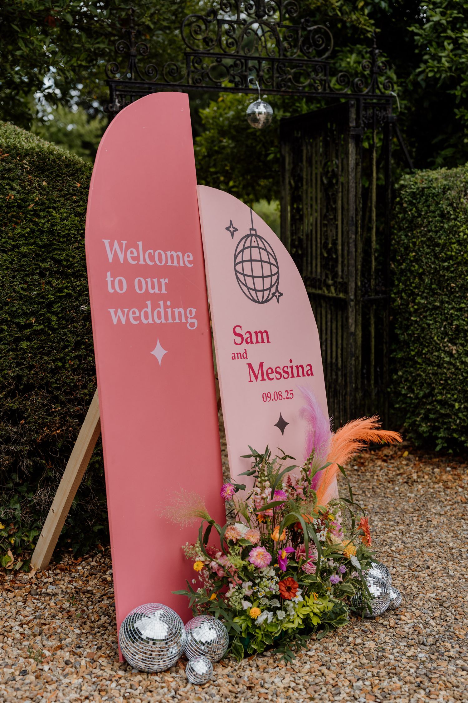 Pink surfboard-style wedding welcome sign with floral decorations and disco balls displayed in front of garden hedges.