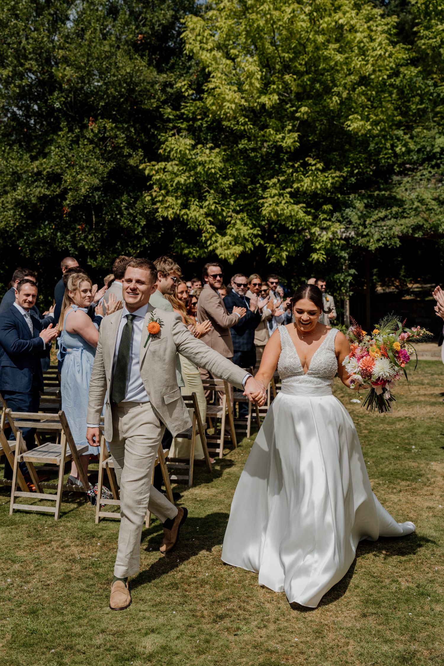 Outdoor wedding ceremony with guests on wooden chairs and lush green foliage in background.