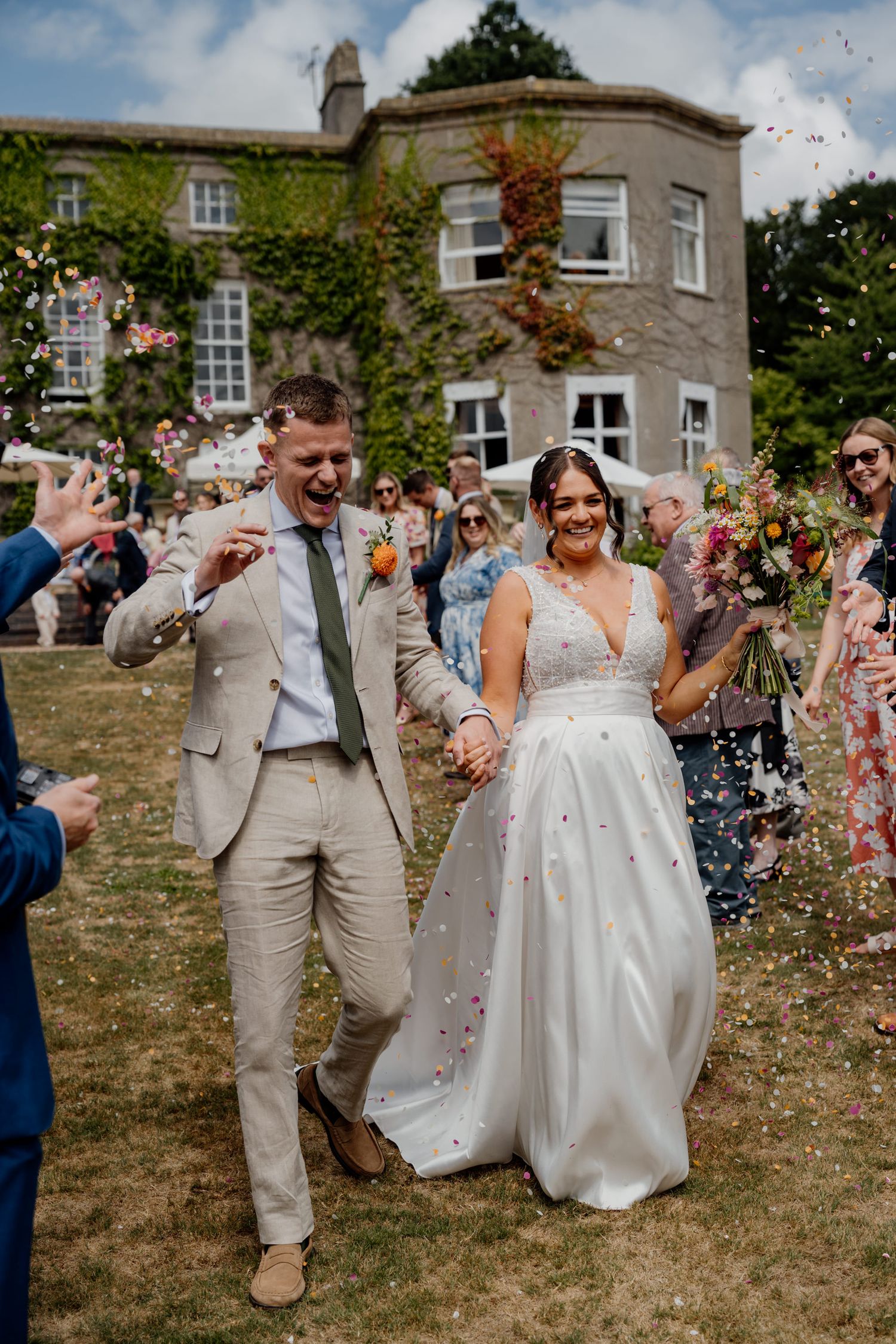 Wedding celebration with guests throwing confetti at a couple in front of an ivy-covered stone building.