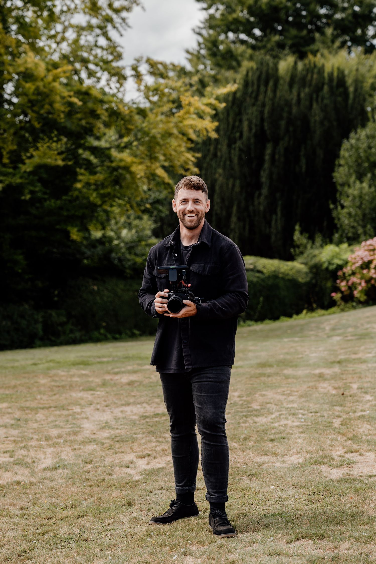 Photographer in black attire laughing while taking pictures at outdoor event.