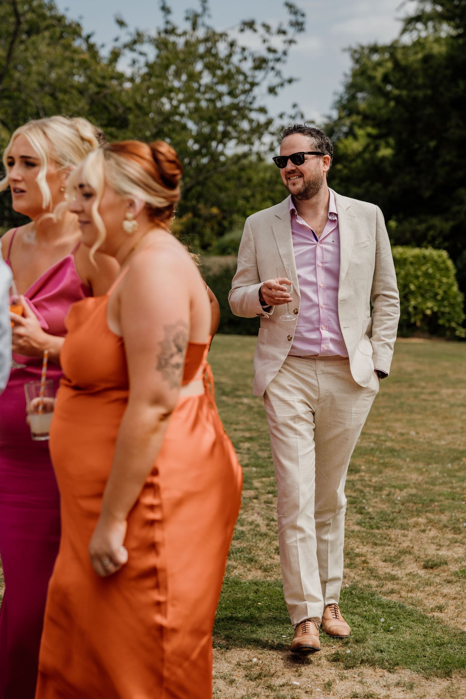 Wedding party in light beige suits and colorful dresses walking outdoors on a sunny day.