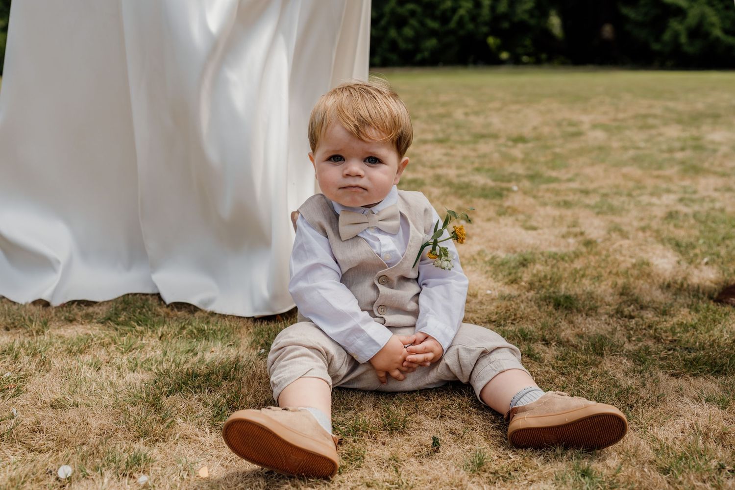 A small child in formal attire sits on grass during an outdoor wedding.
