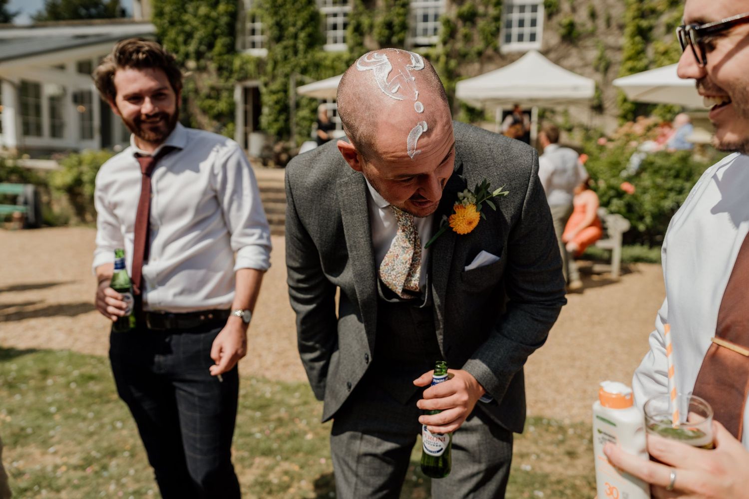 Group in formal attire sharing drinks and laughter outdoors at a garden event on a sunny day.