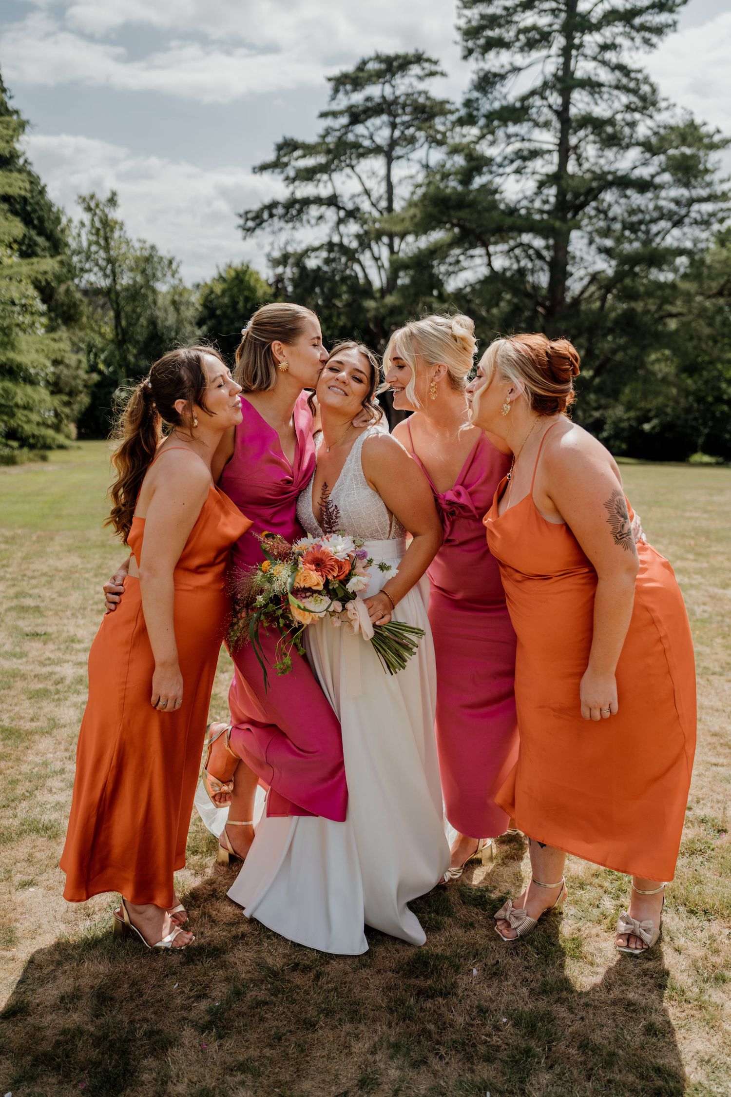 Bridesmaids in orange and pink dresses pose together on a grassy hill.
