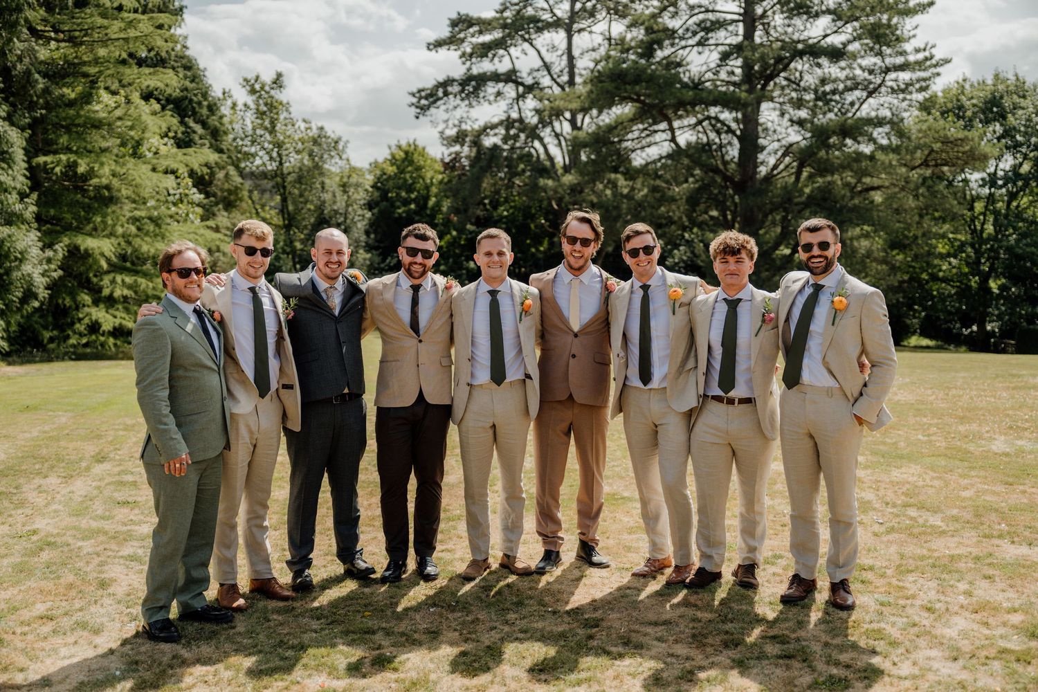 A group of groomsmen in light colored suits pose together outdoors on a sunny day with trees in the background.
