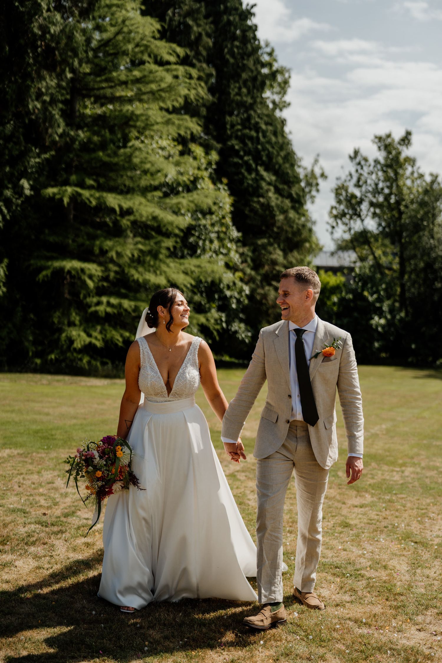 A couple in wedding attire walk together on a sunny grassy field with trees in the background.