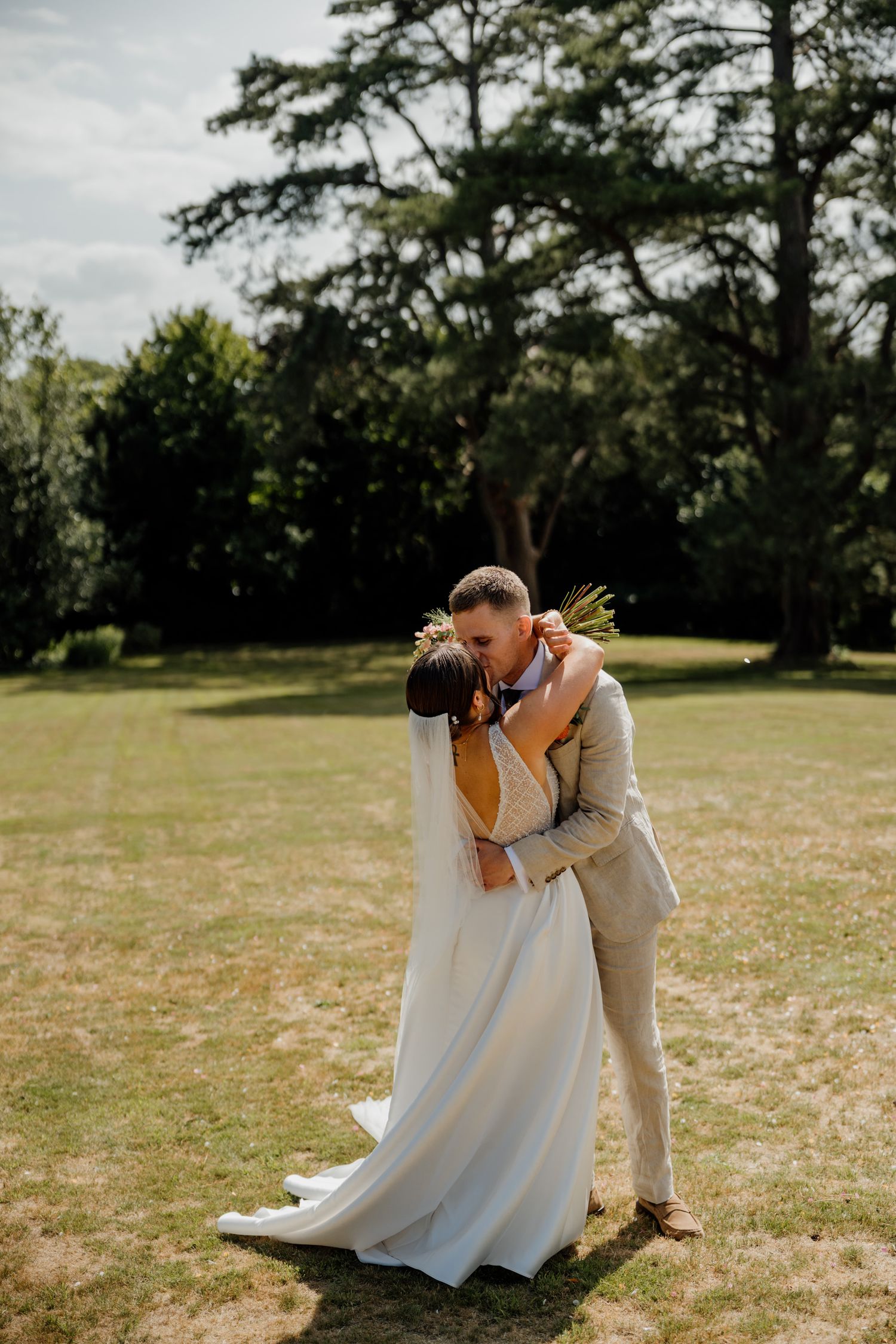 A couple shares a romantic kiss in an outdoor setting with a dreamy sunlit field and tall trees behind them.