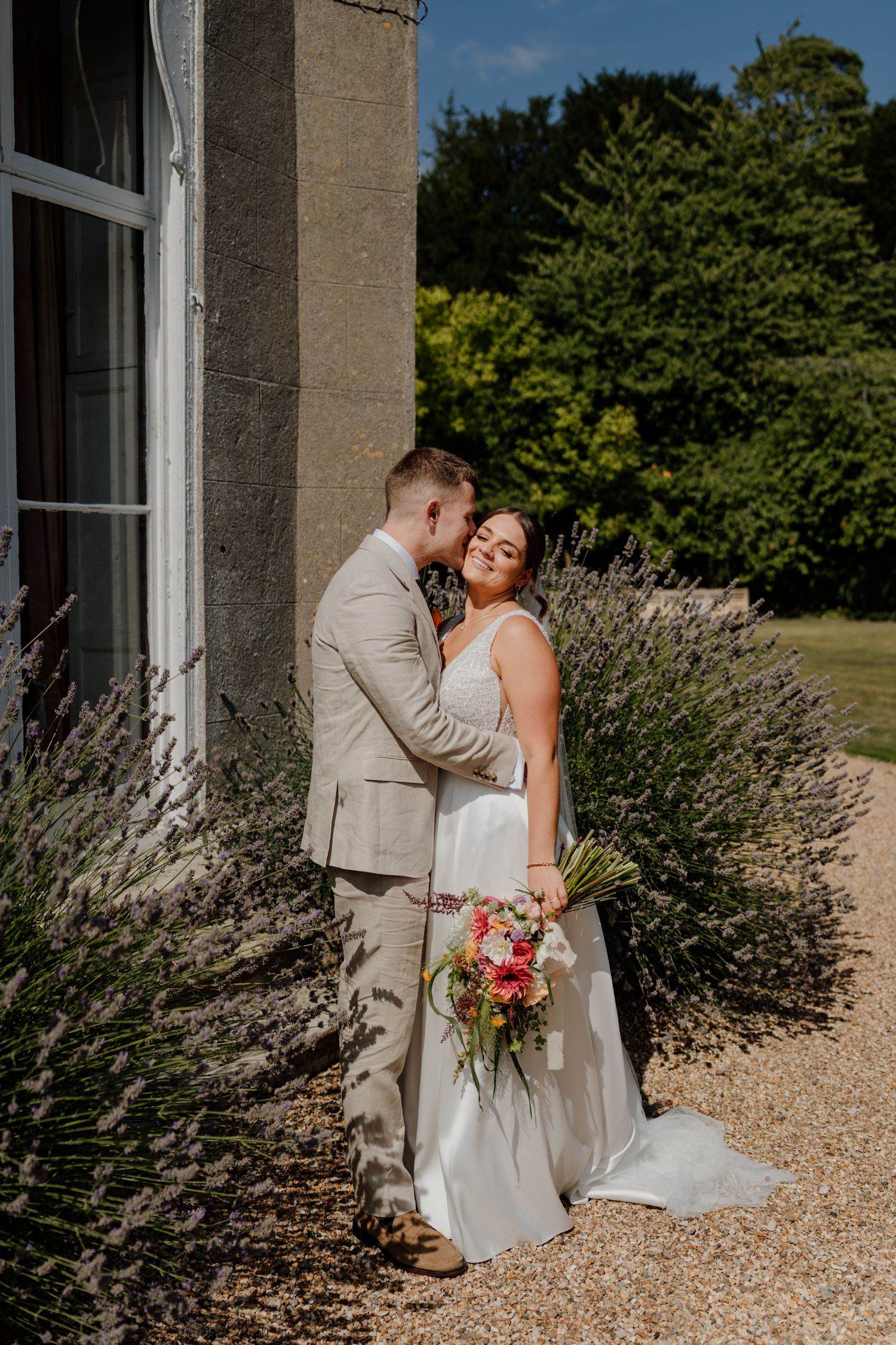 A romantic wedding couple shares intimate moments against a stone building backdrop with lavender bushes.