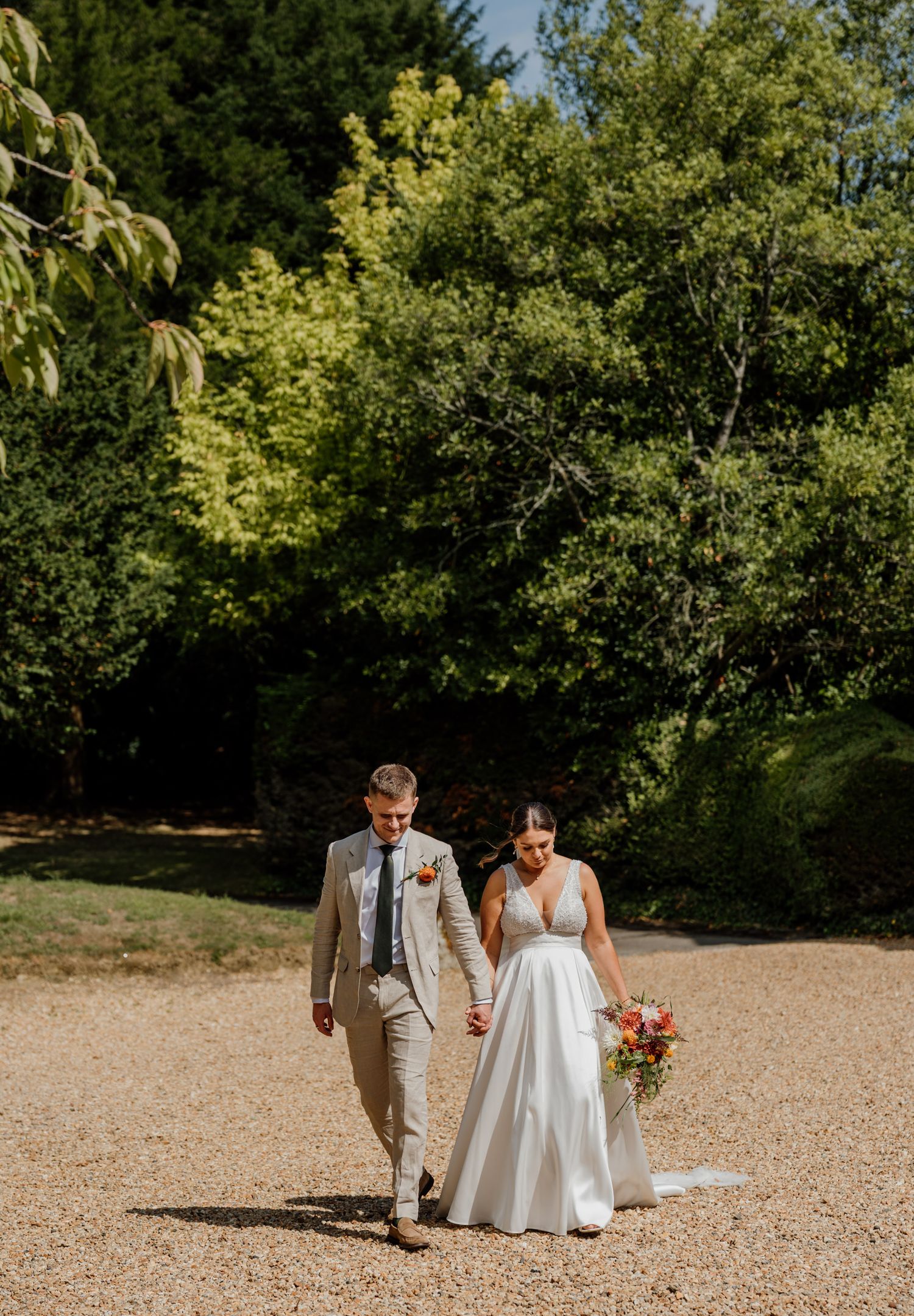 A wedding couple walks together on a gravel path surrounded by lush green trees.