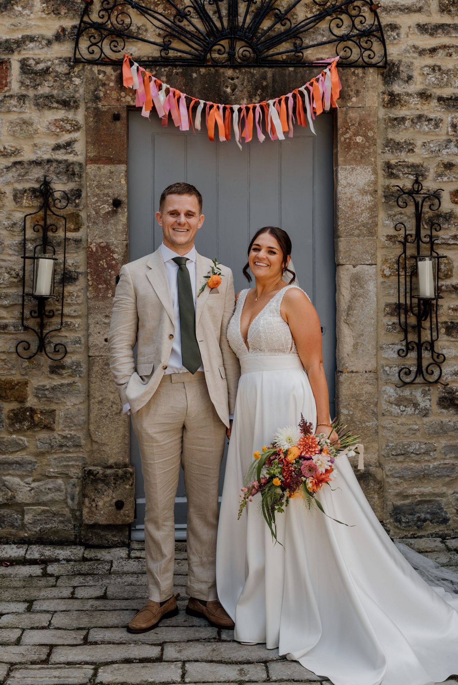 A wedding couple poses in front of a rustic stone wall with ornate iron details and colorful bunting overhead.