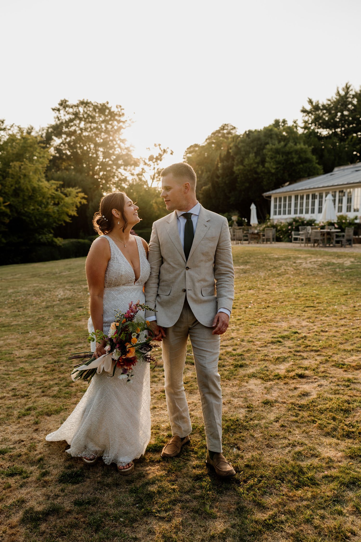 Newlyweds pose for sunset photos in a grassy field with a white building in the background.
