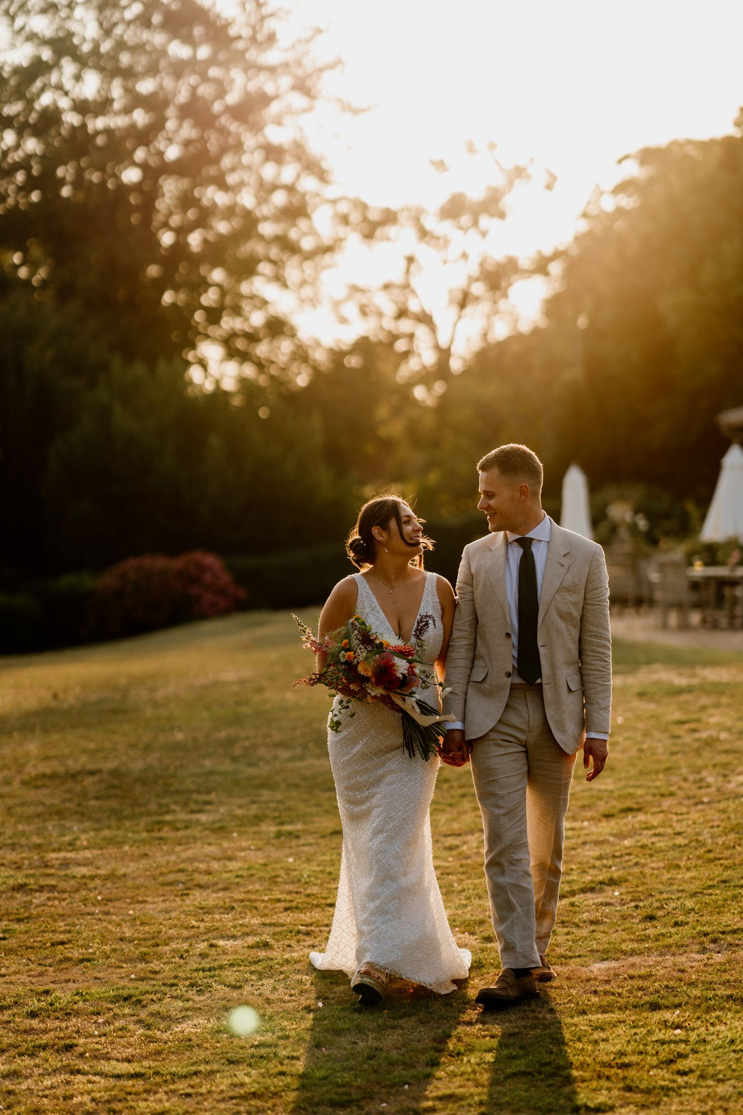 A newlywed couple walking through a golden-lit field at sunset during their intimate outdoor wedding.